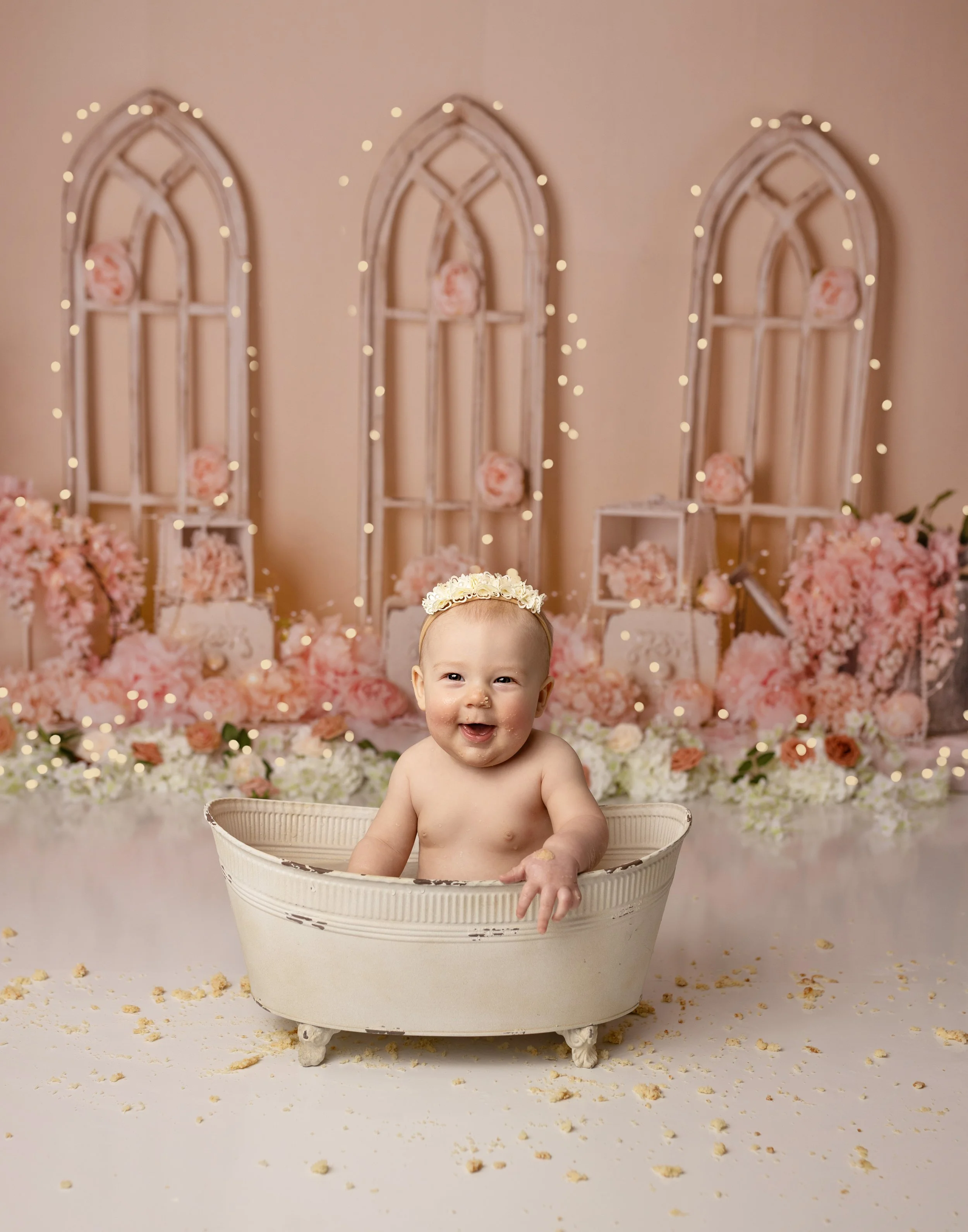 A smiling baby sitting in a vintage white tub during a celebration, with pink and white flowers and soft lights in the background.