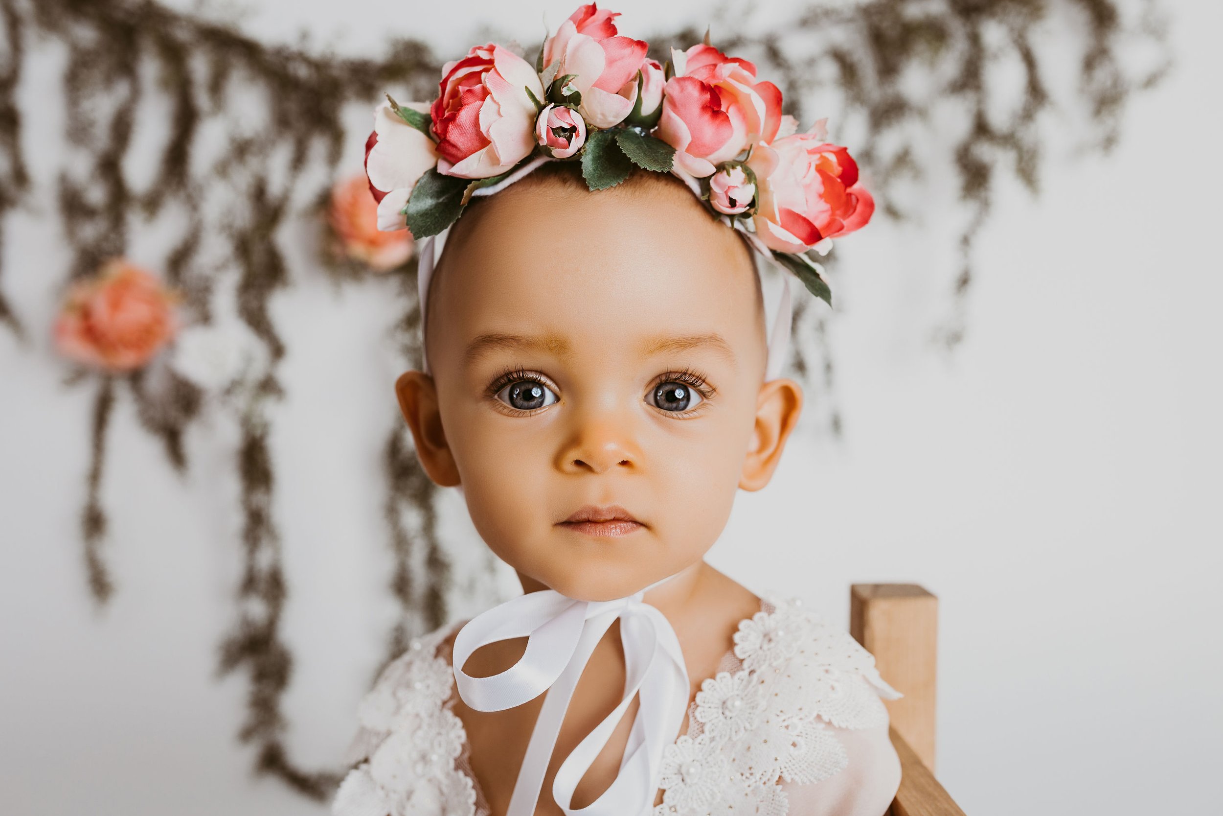Baby wearing floral headband and white lace dress with floral backdrop.