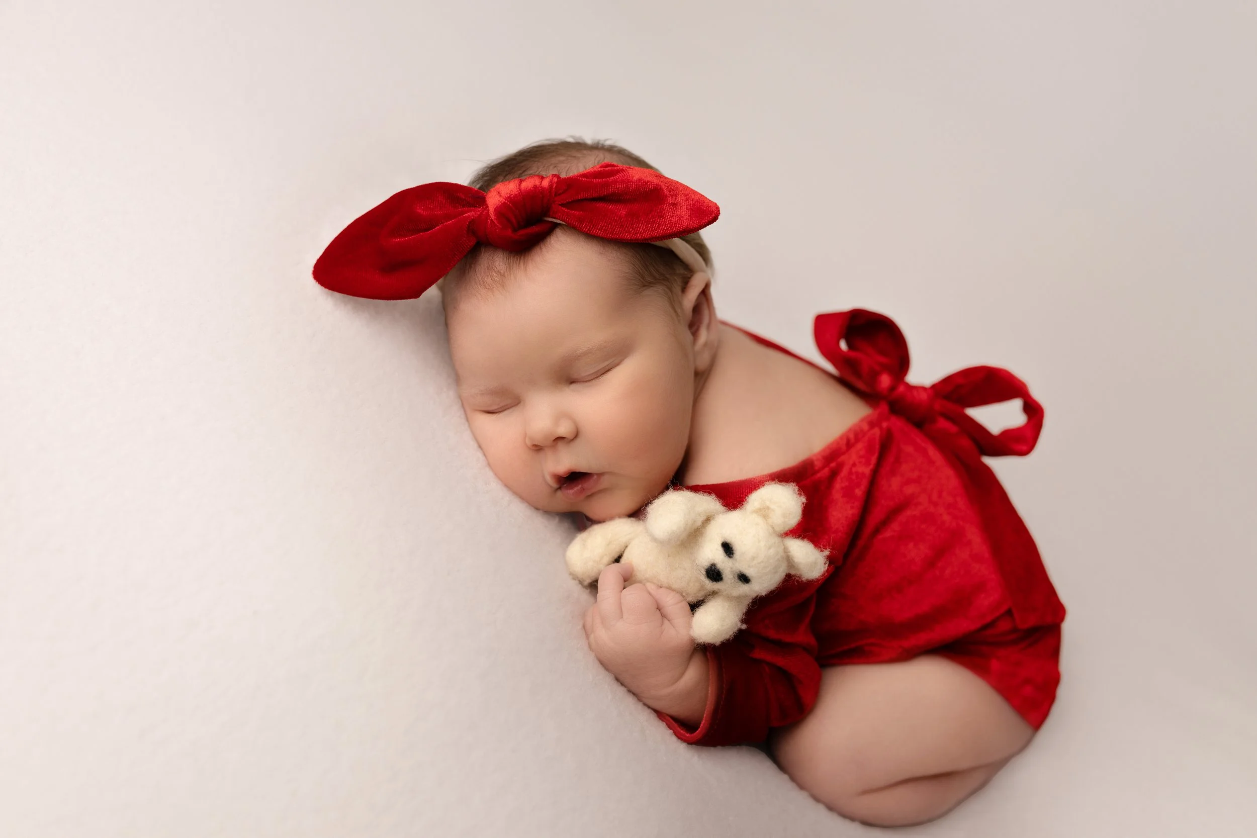 A sleeping baby girl wearing a red headband and red outfit, holding a small plush dog against a white background.