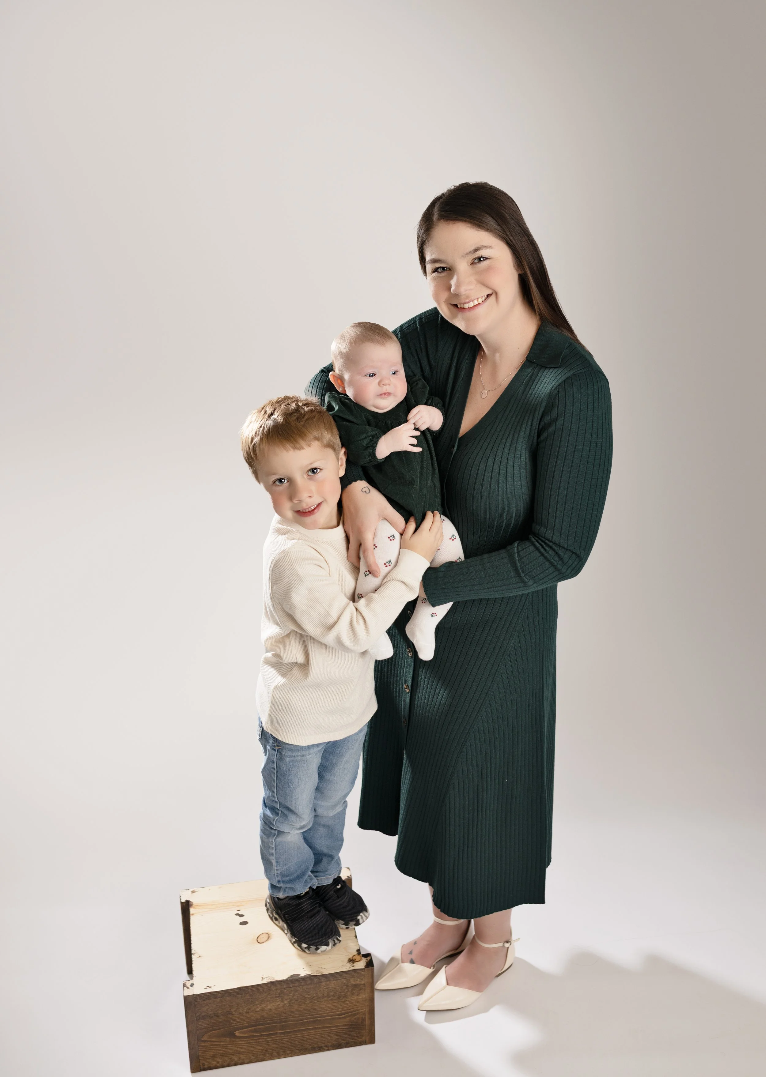 A woman with long dark hair, wearing a dark green ribbed dress and beige heels, holding a baby girl in her right arm. A young boy with light brown hair, wearing a cream sweater, blue jeans, and black sneakers, standing on a wooden box and holding the