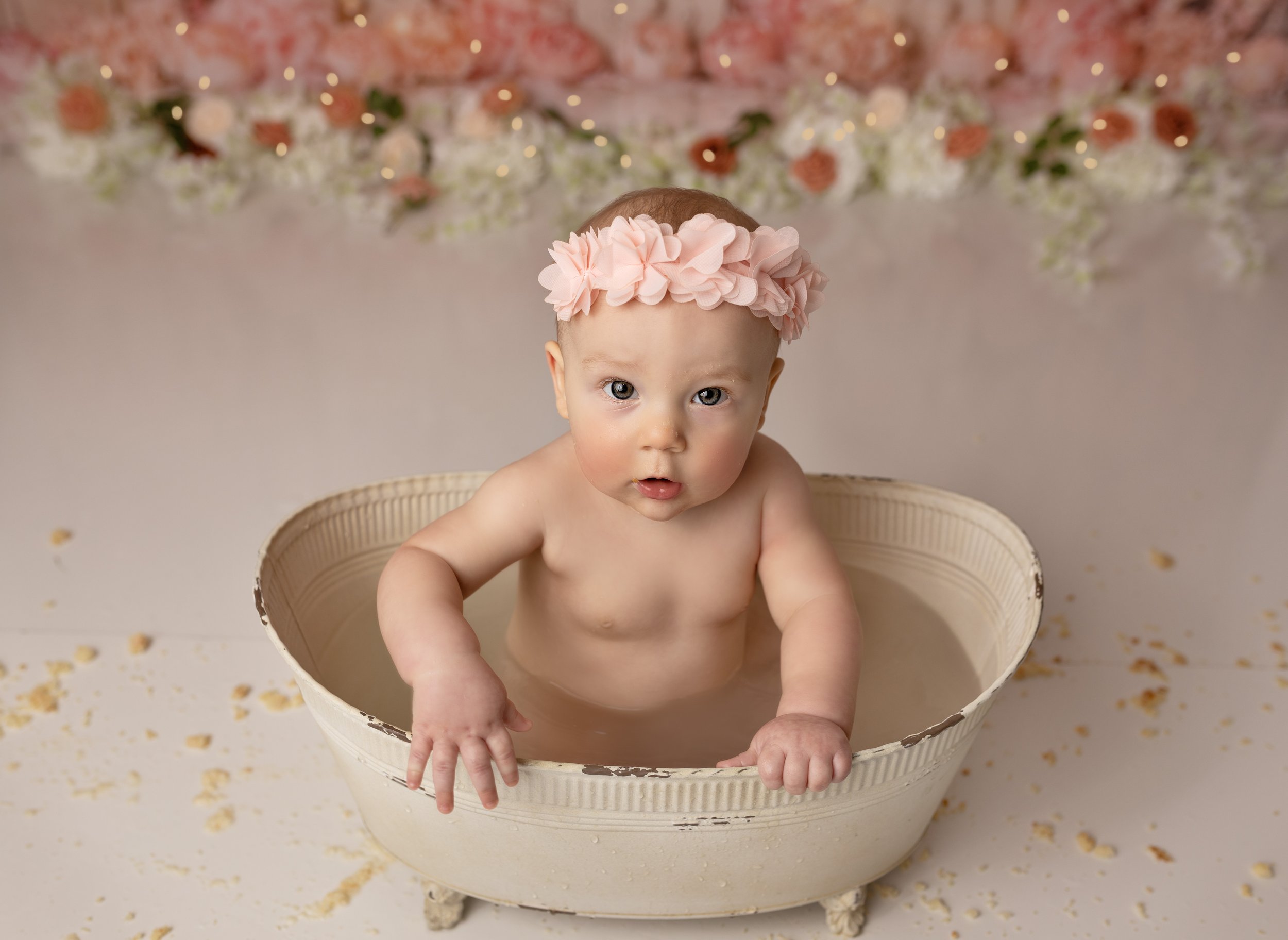 Baby sitting in a vintage bathtub, wearing a pink flower headband, with a floral backdrop and scattered yellow flower petals on the floor.