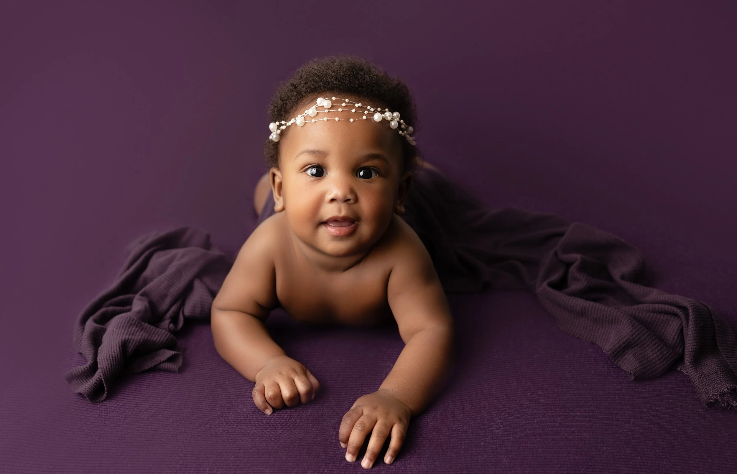 A baby laying on a purple blanket, wearing a pearl headband, looking at the camera.