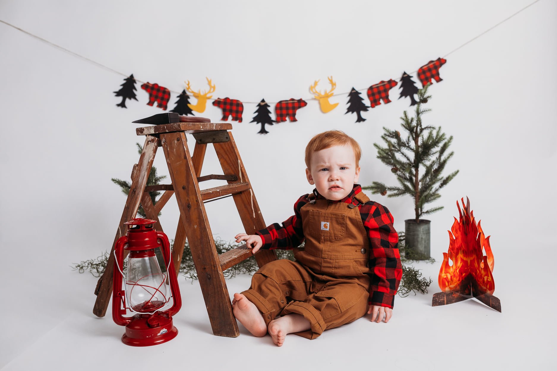 Toddler in brown overalls and plaid shirt sitting next to a wooden step ladder with a red lantern, in a rustic themed setting with Christmas-style decorations, pine trees, and a faux campfire.