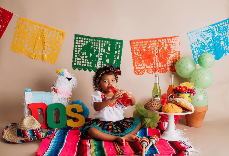 A child wearing festive clothing sitting on a colorful blanket with traditional Mexican decorations. There are papel picado banners, a piñata, the word 'DOS,' and a straw hat. The child is drinking from a bottle, surrounded by Mexican pastries, marac