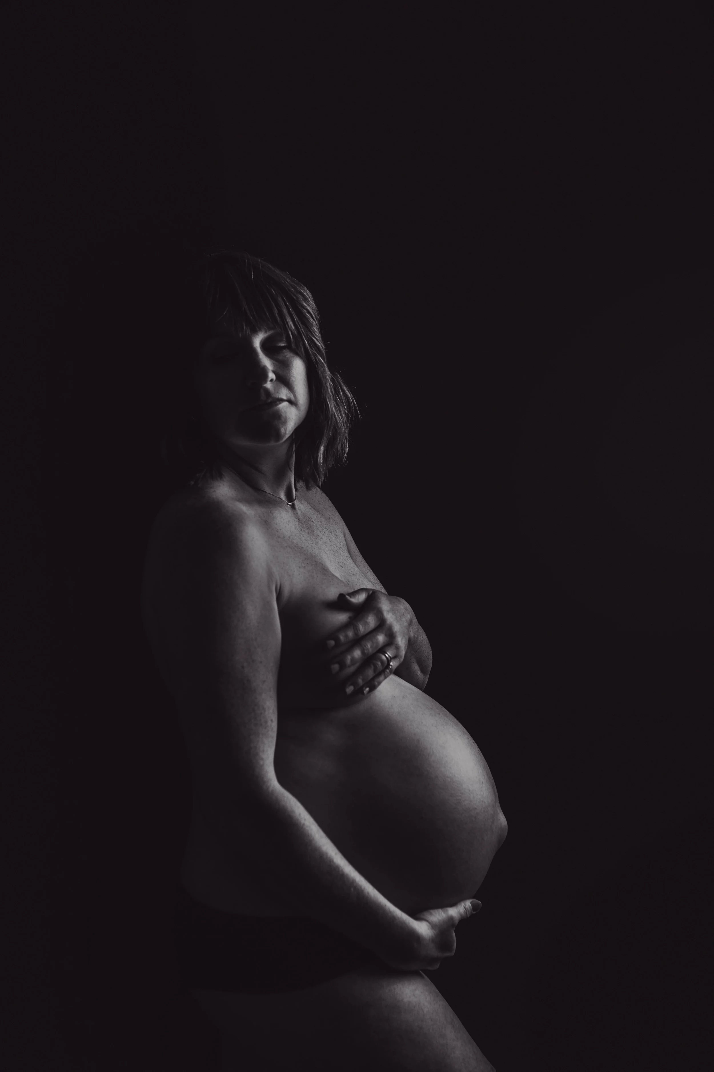 Black and white photograph of a pregnant woman holding her belly with one hand and covering her breast with the other, standing against a dark background. Louisville ky maternity photographer