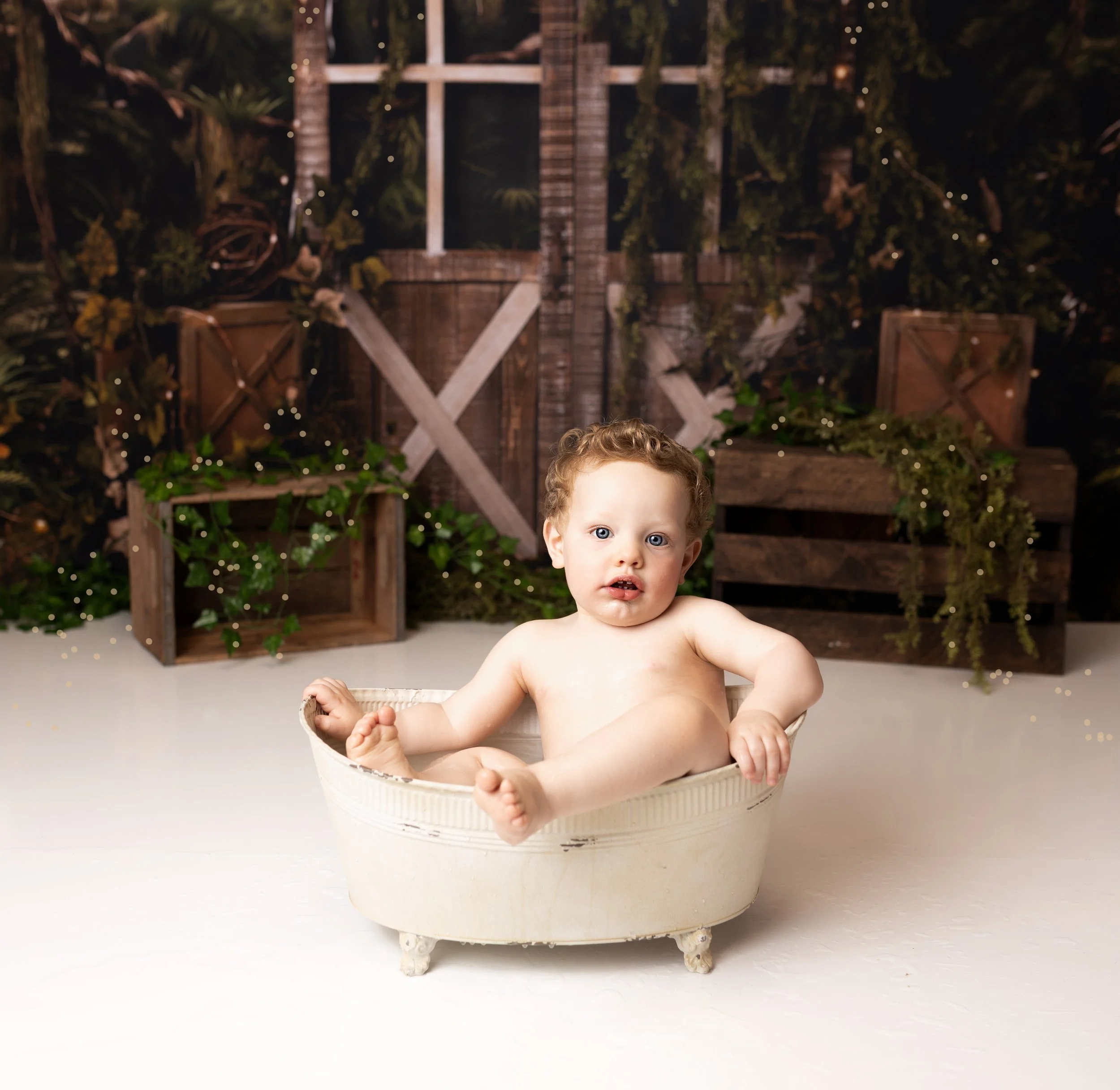A young child with curly red hair sitting in a vintage white bathtub, surrounded by a rustic woodland backdrop with greenery and fairy lights.