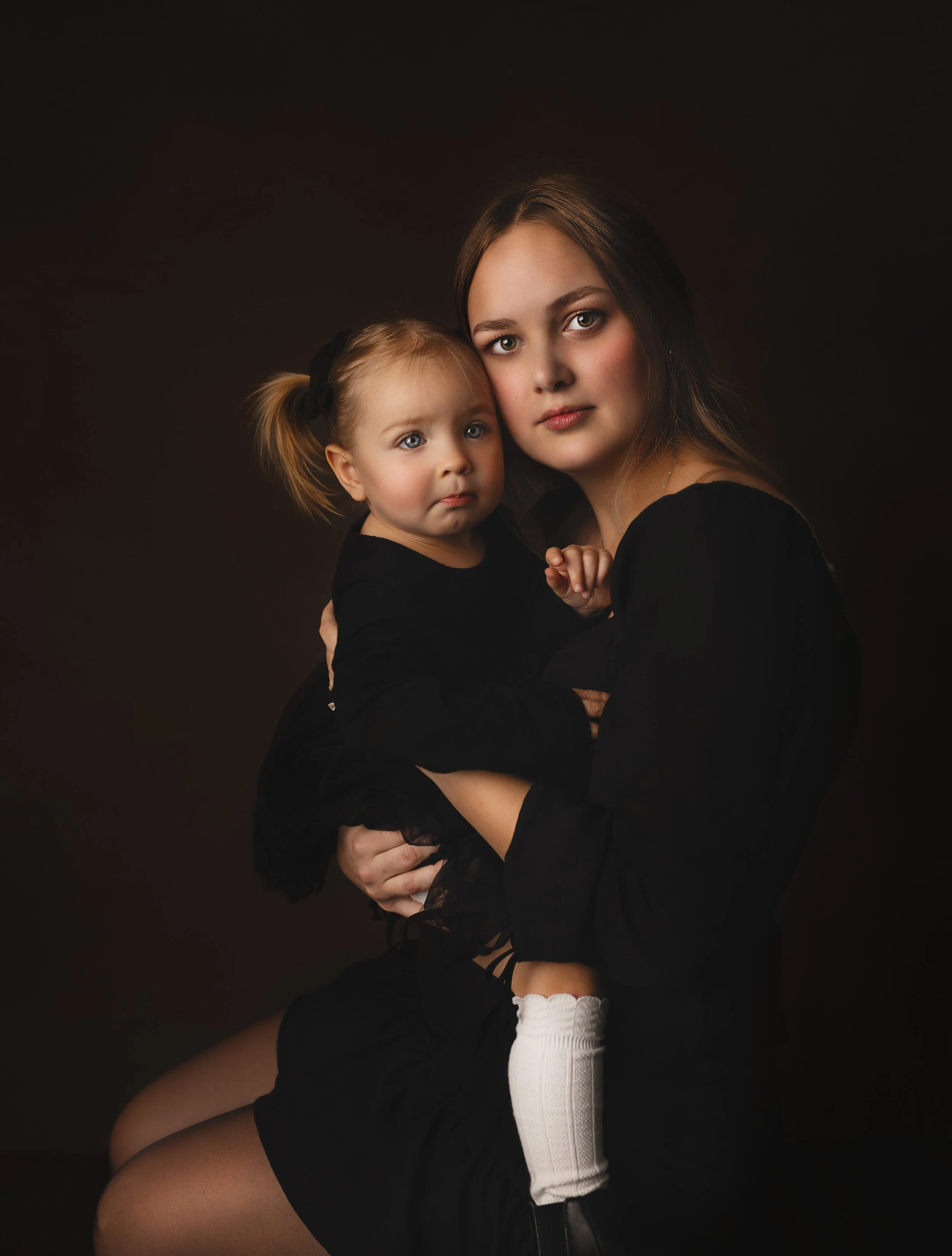 A woman holding a young girl against a dark background, both dressed in black. The woman has long brown hair, and the girl has pigtails secured with black bows. The girl is wearing white knee-high socks.