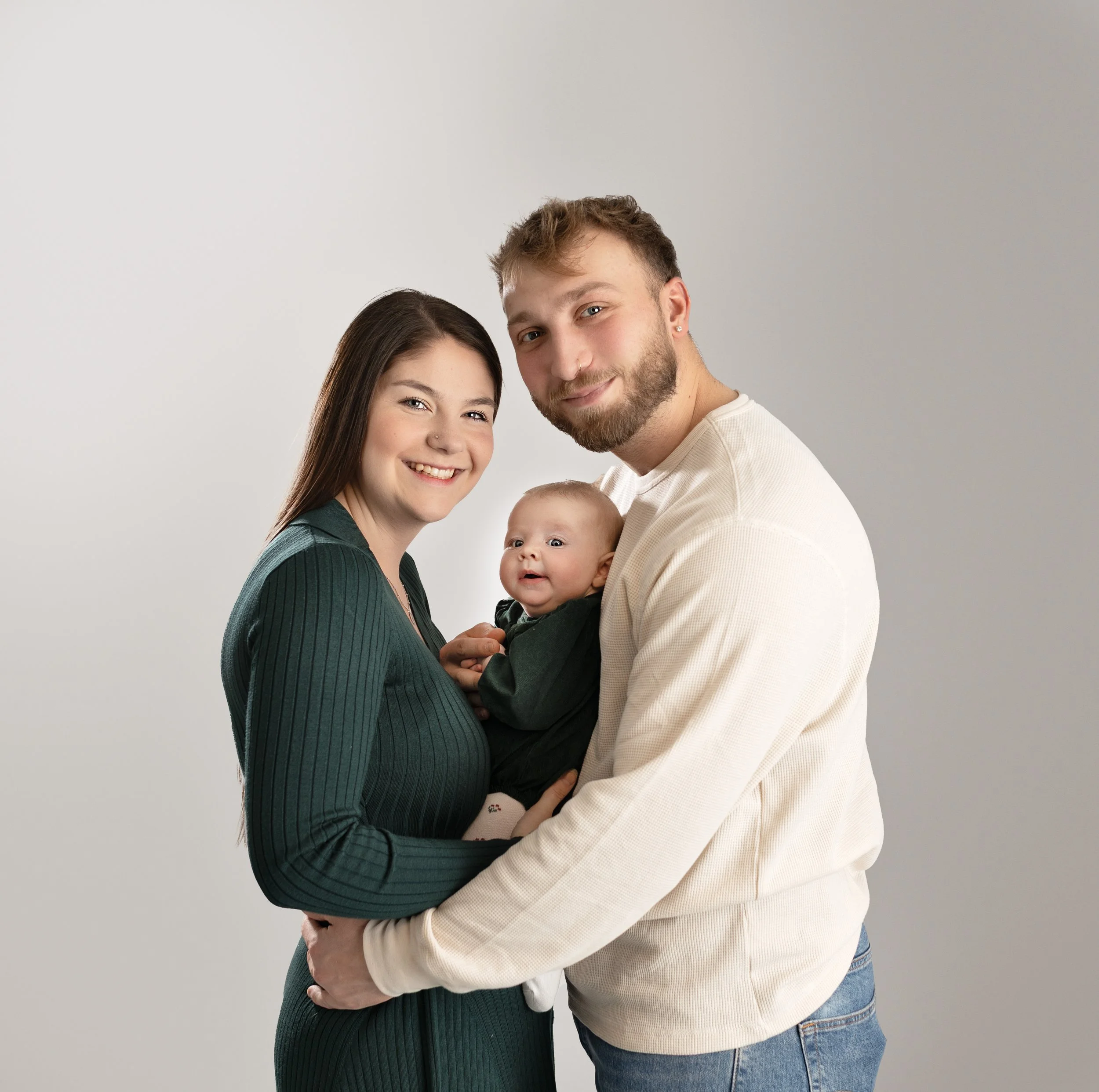 A happy family of three: a woman, a man, and a baby, standing close together and smiling against a plain white background.