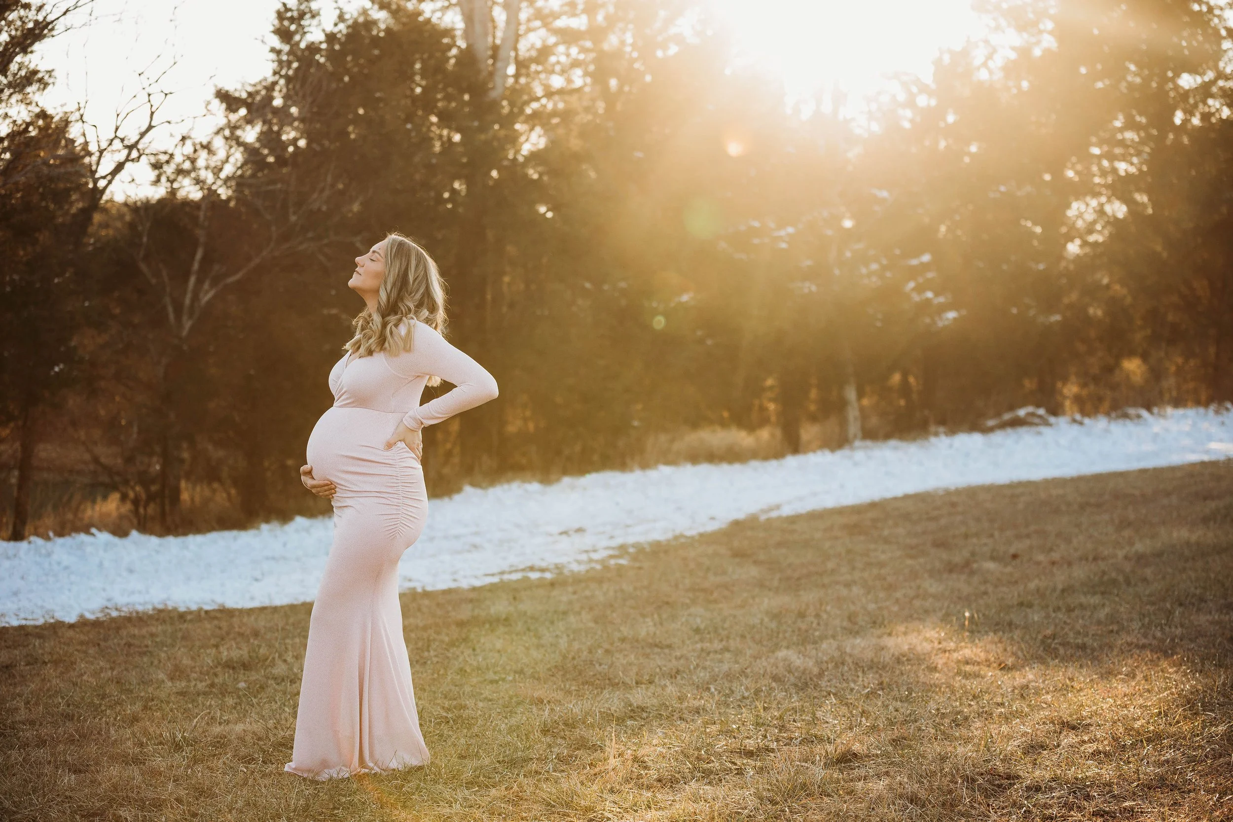 Pregnant woman in a light pink dress standing outdoors during sunset, with her eyes closed and hands on her pregnant belly. Louisville ky maternity photographer
