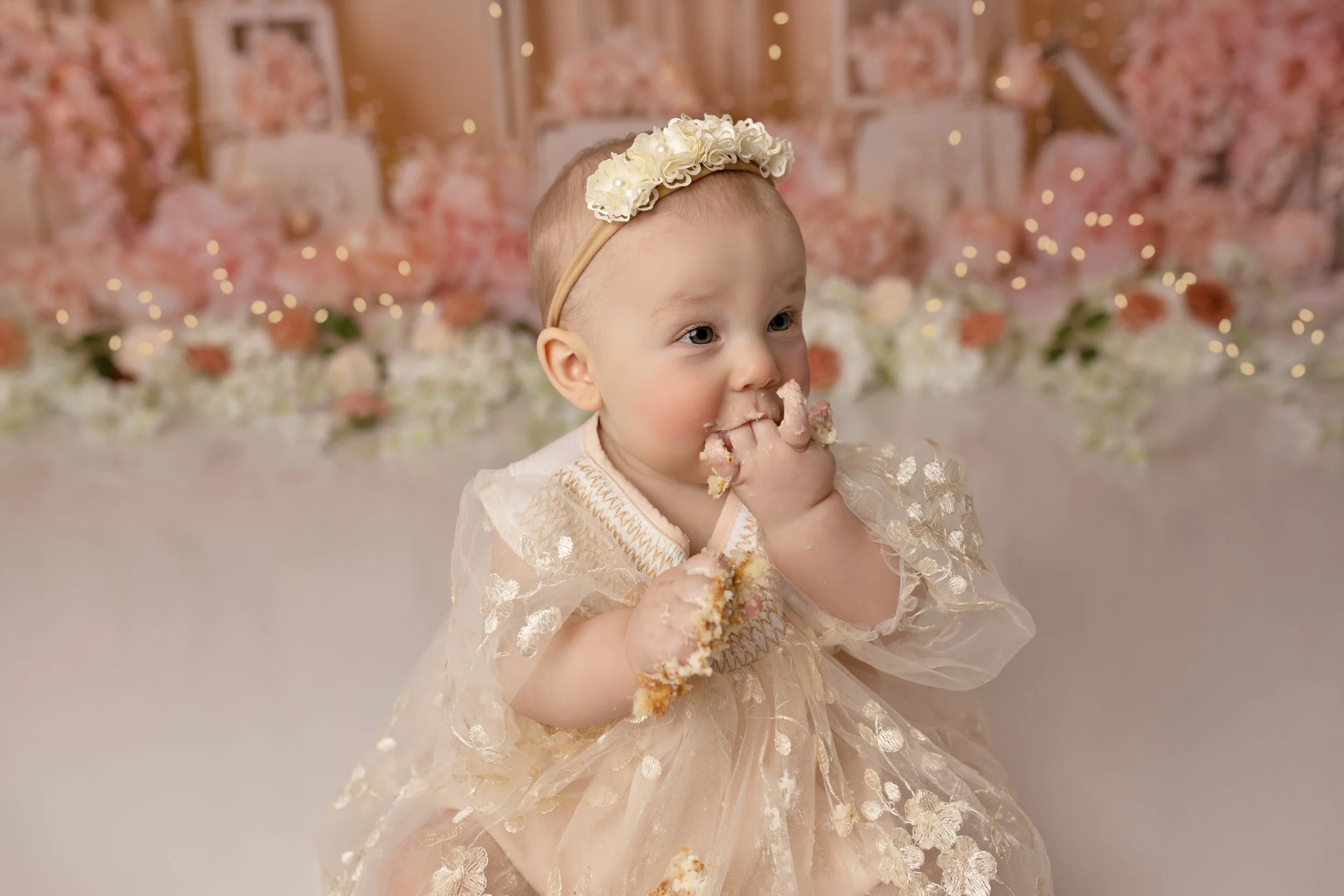 A baby girl dressed in a fancy dress with a floral headband eating cake at a celebration with pink and white flowers and fairy lights in the background.