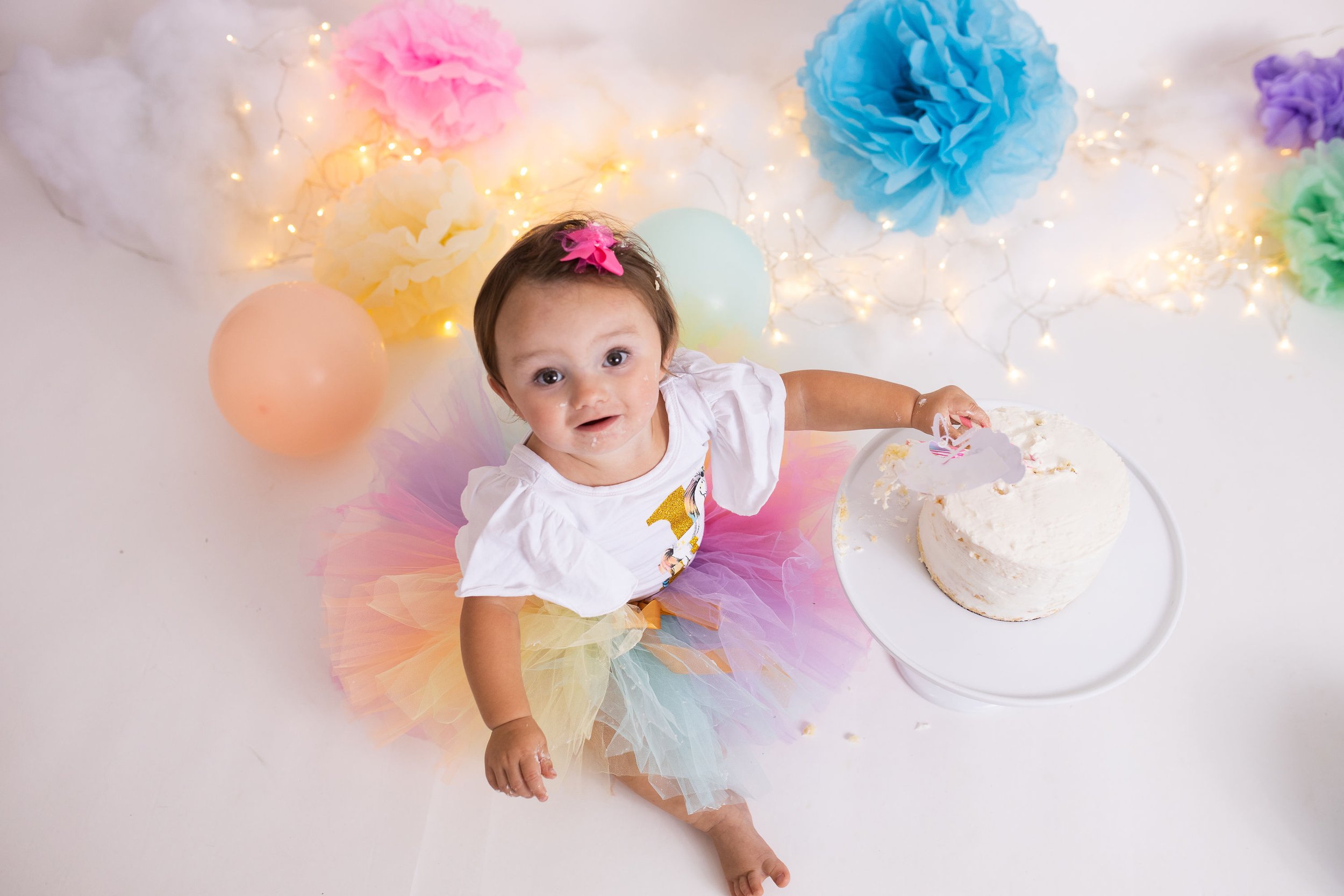 Baby in colorful tutu with cake, surrounded by balloons and paper flowers.