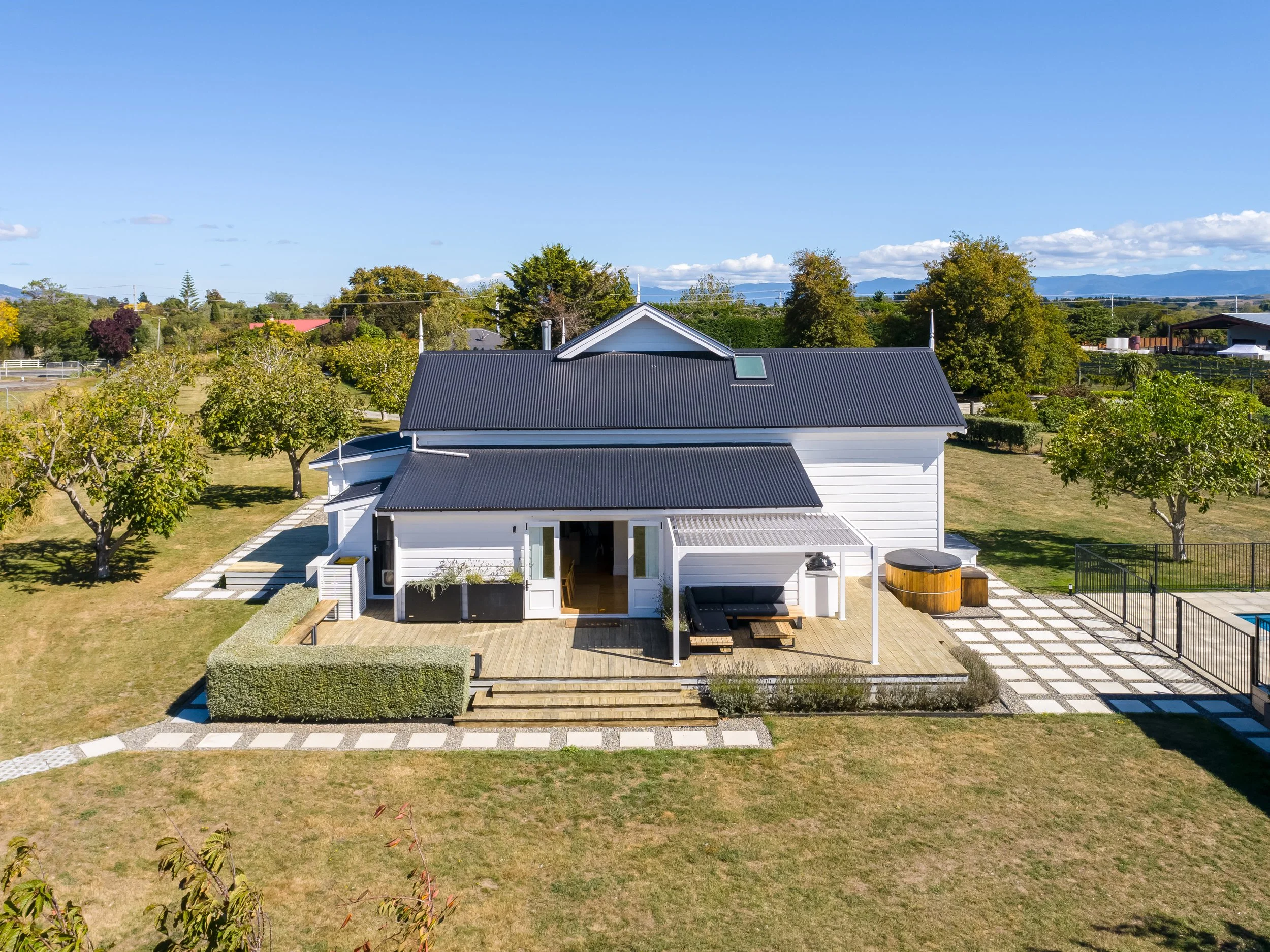 Aerial view of a large, charming white Victorian villa with a big sunny back deck area that has couches under a shaded pergola, along with an outdoor kitchen and spa, surrounded by well-kept lawns and trees under a blue sky.