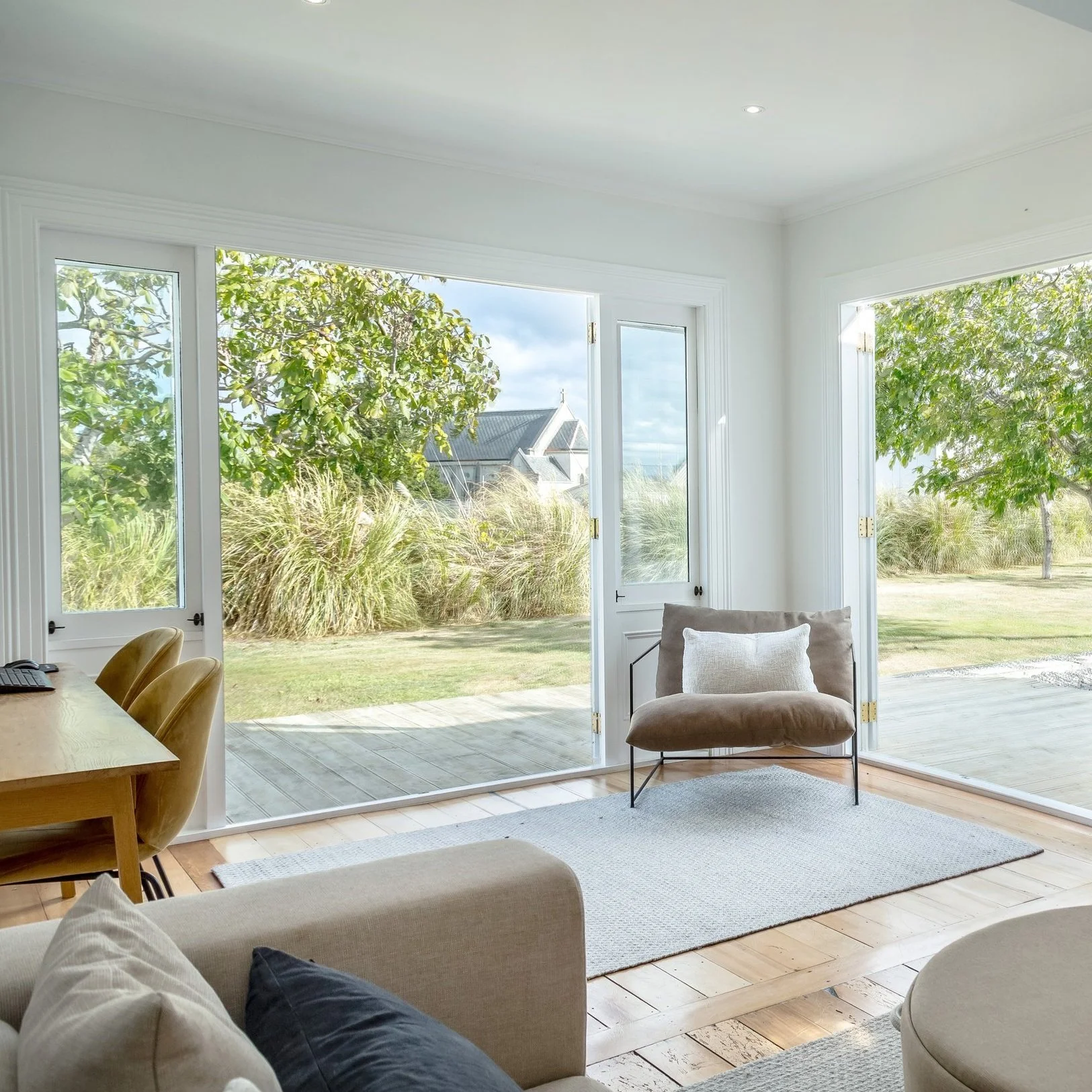 A bright living room with simple white walls and light-coloured wood floor, leading to french doors that open onto a green garden with a beautiful church in the background. Great for weekend stays, small weddings, post wedding events and groups.
