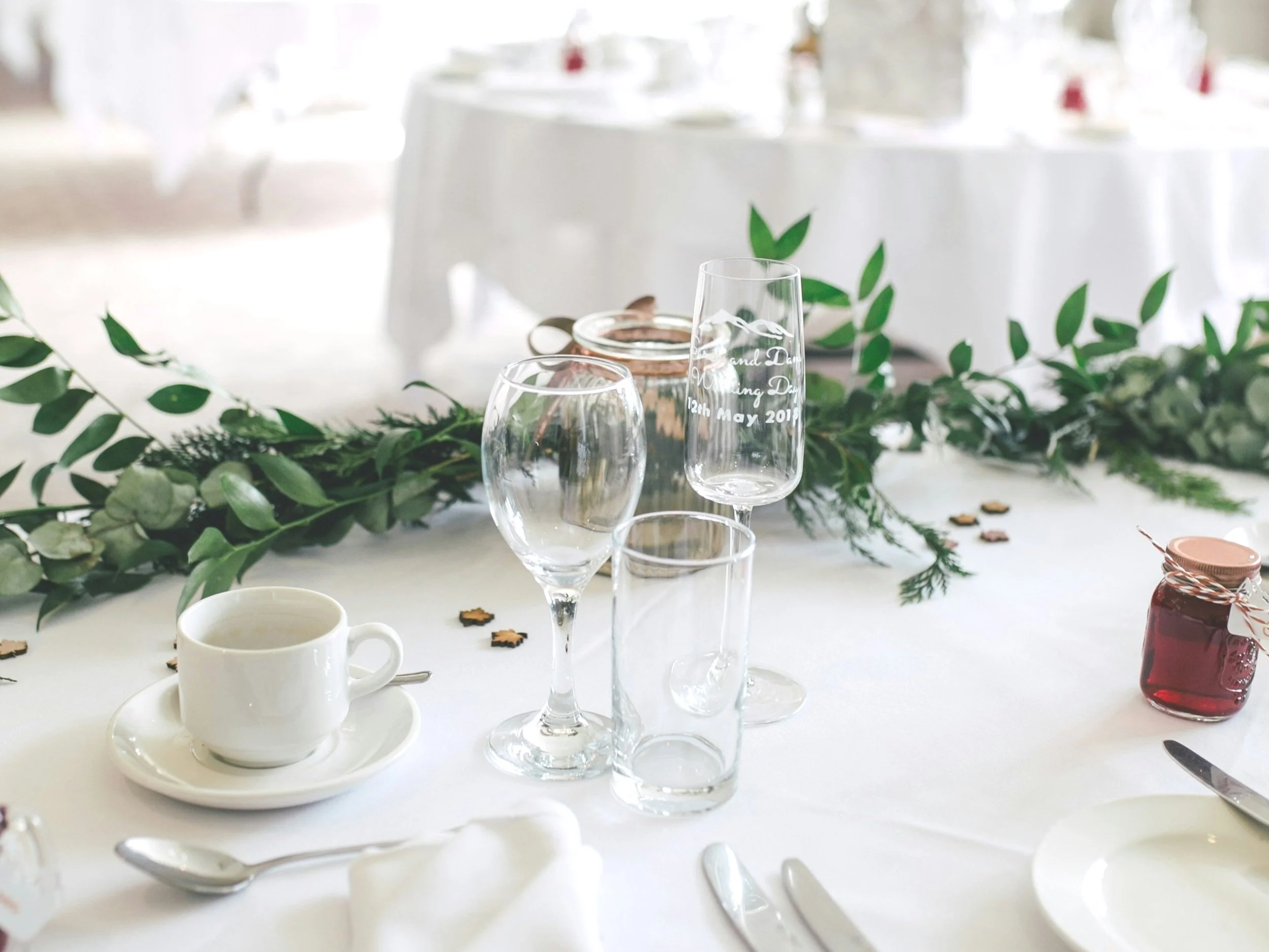 Simple white tables set for a wedding or celebration with sparkling wine glasses, white plates and cups, elegant silver and decorative green leafy garlands. Great for small weddings, post wedding events, group get-togethers and celebrations.