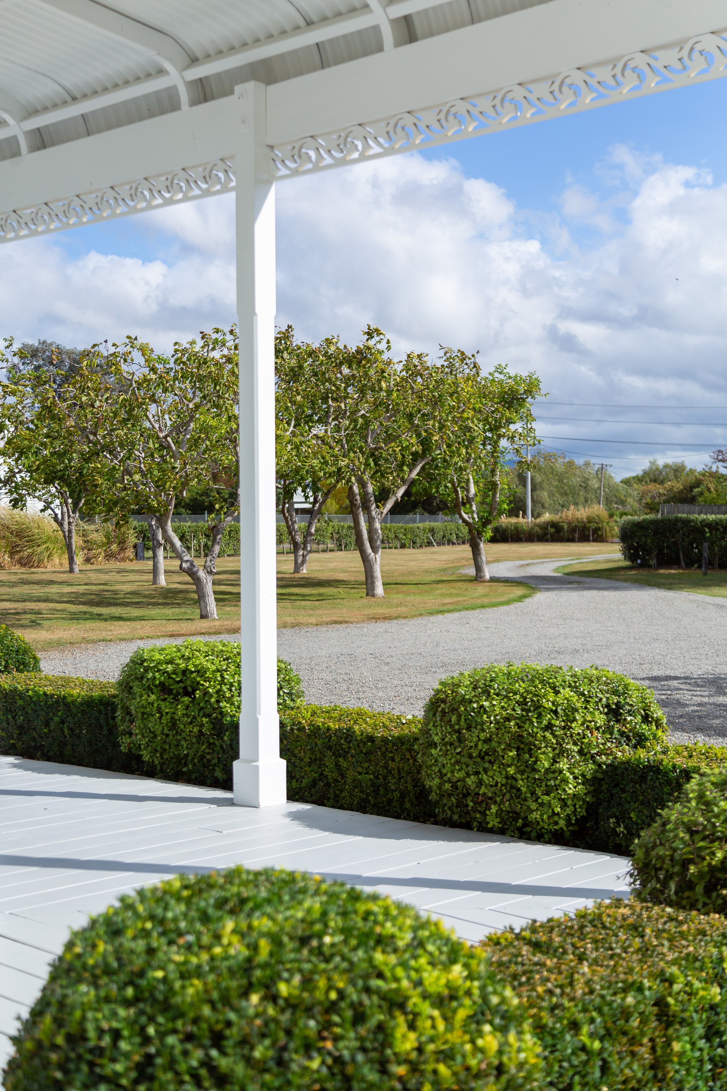A white porch with decorative trim, surrounded by topiary looking out to trees and a curving stone driveway. Great for weekend stays, small weddings, post wedding events, group gatherings, celebrations and holidays.