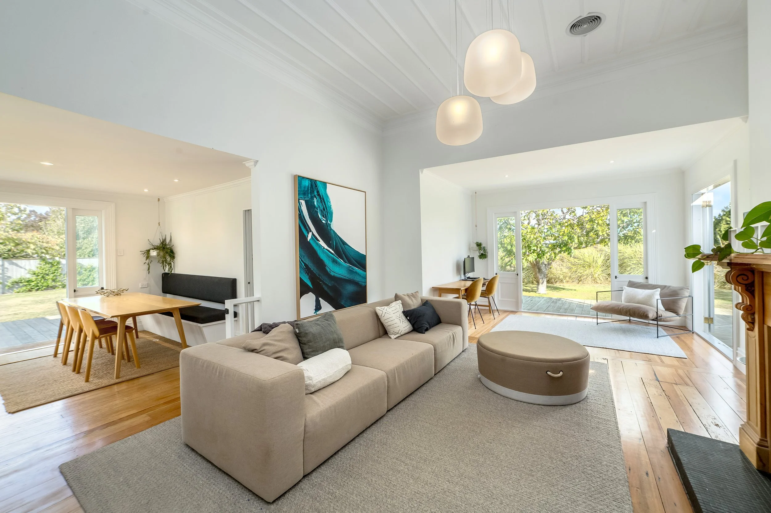 A bright white, contemporary living room that has a large sofa and ottoman, light wooden floor and modern decor, with french doors opening to a lush outdoor garden. Great for weekend stays, group gatherings, celebrations and holidays.