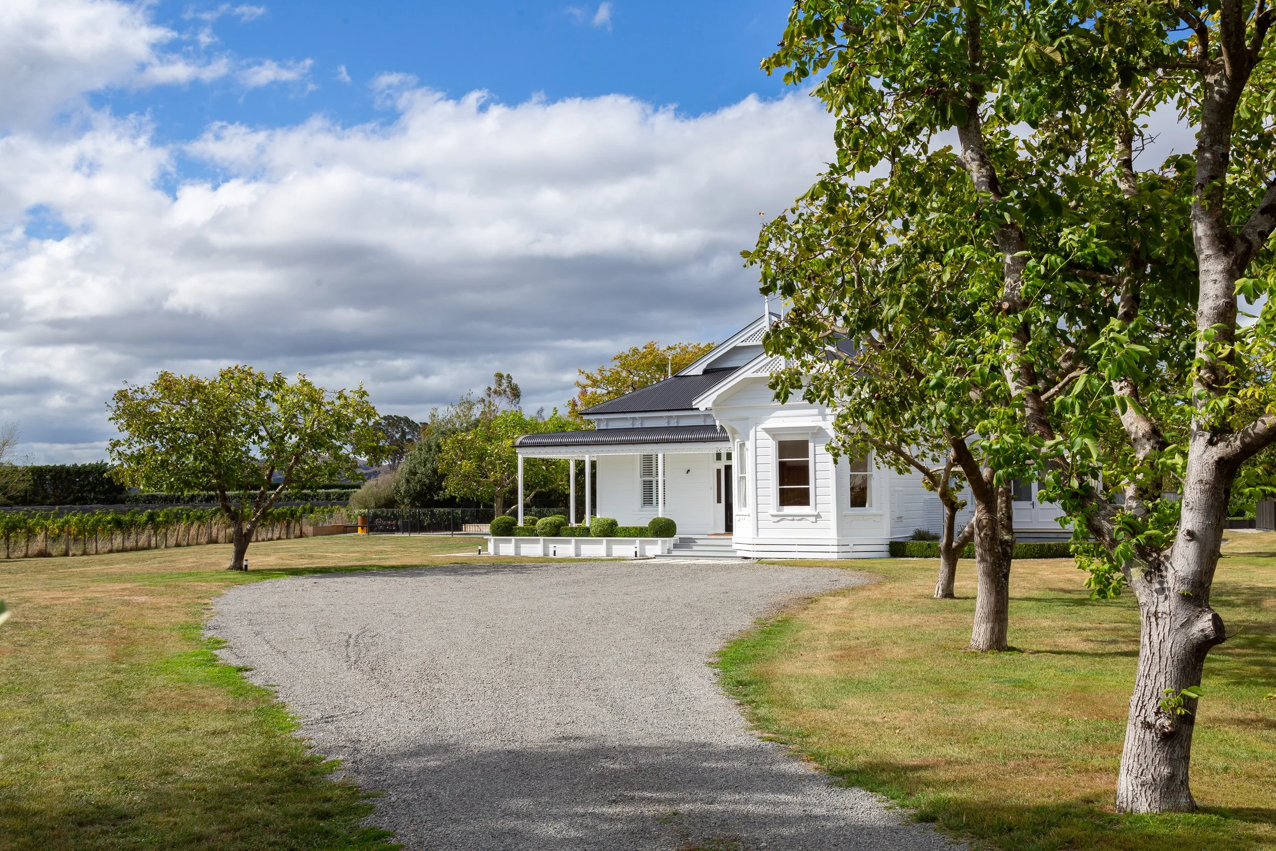 A stone driveway curving through a grassy lawn to a large, charming white Victorian villa with a rolled verandah over the wrap-around porch, surrounded by trees and set against a blue sky dotted with a few white clouds.