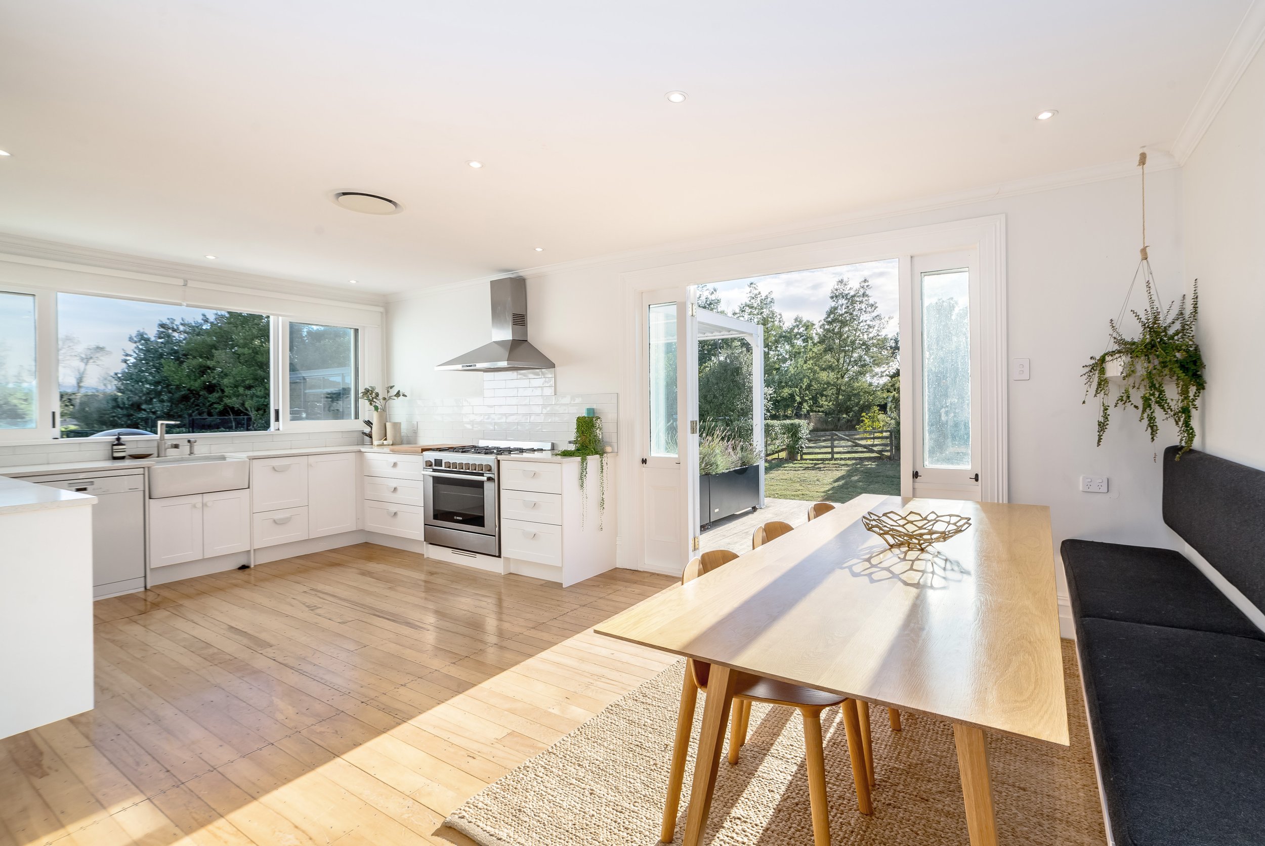 A bright white kitchen with warm sun streaming into it over light wood floors and wooden table, with chairs, a seat bench and french doors open, overlooking a wooden deck, green lawn and gardens.