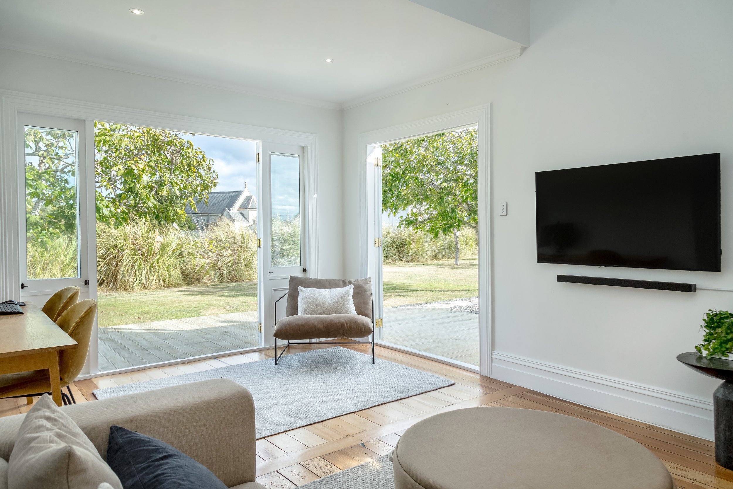 A bright living room with simple white walls and light-coloured wood floor, leading to french doors that open onto a green garden with a beautiful church in the background. Great for weekend stays, group gatherings, celebrations and holidays.
