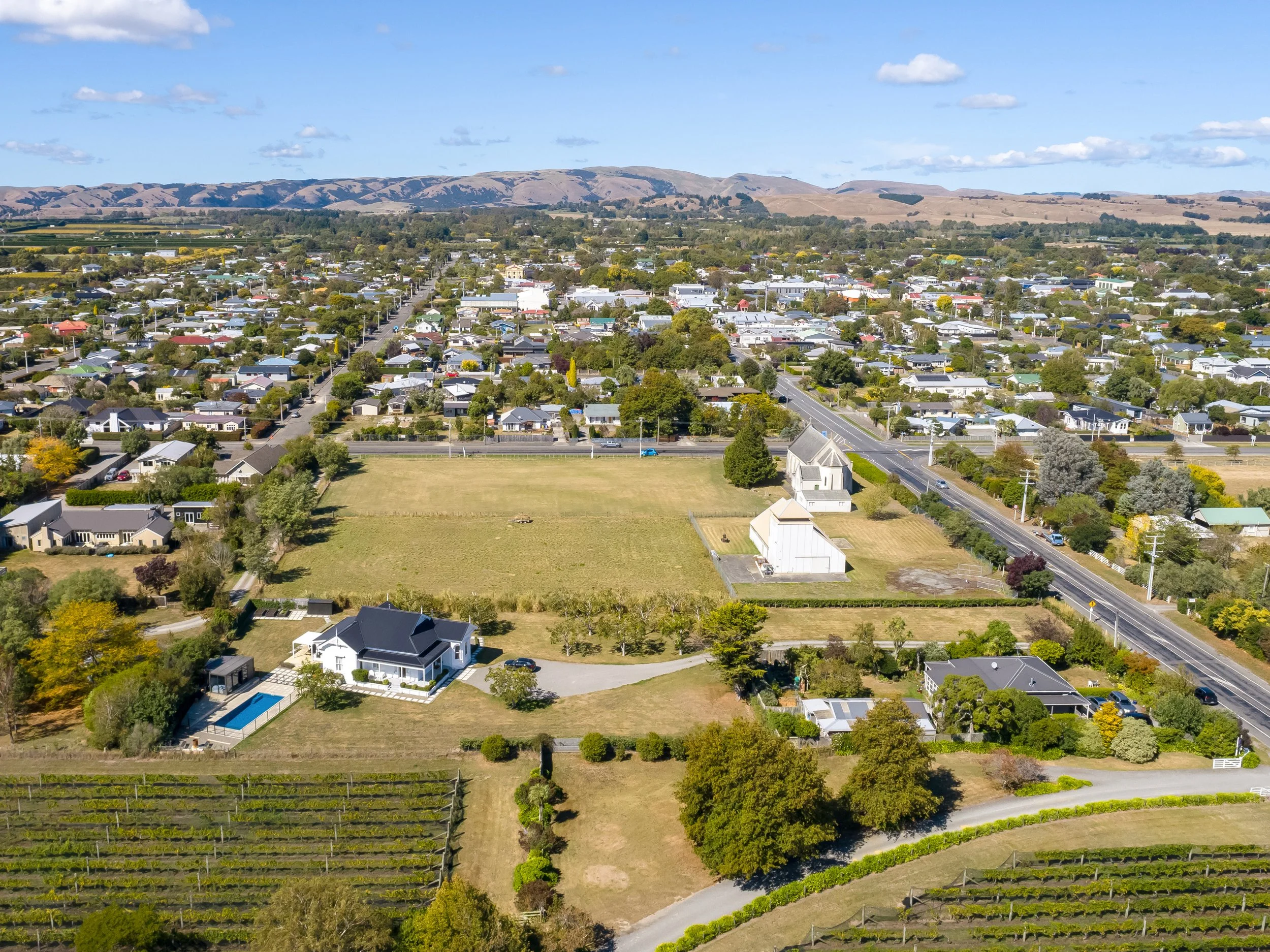 Aerial view of a large white villa and swimming pool on a sprawling country section, set amongst fields and vineyards, near a beautiful church and building, close to the centre of Martinborough township. Great for weekend stays and small weddings.