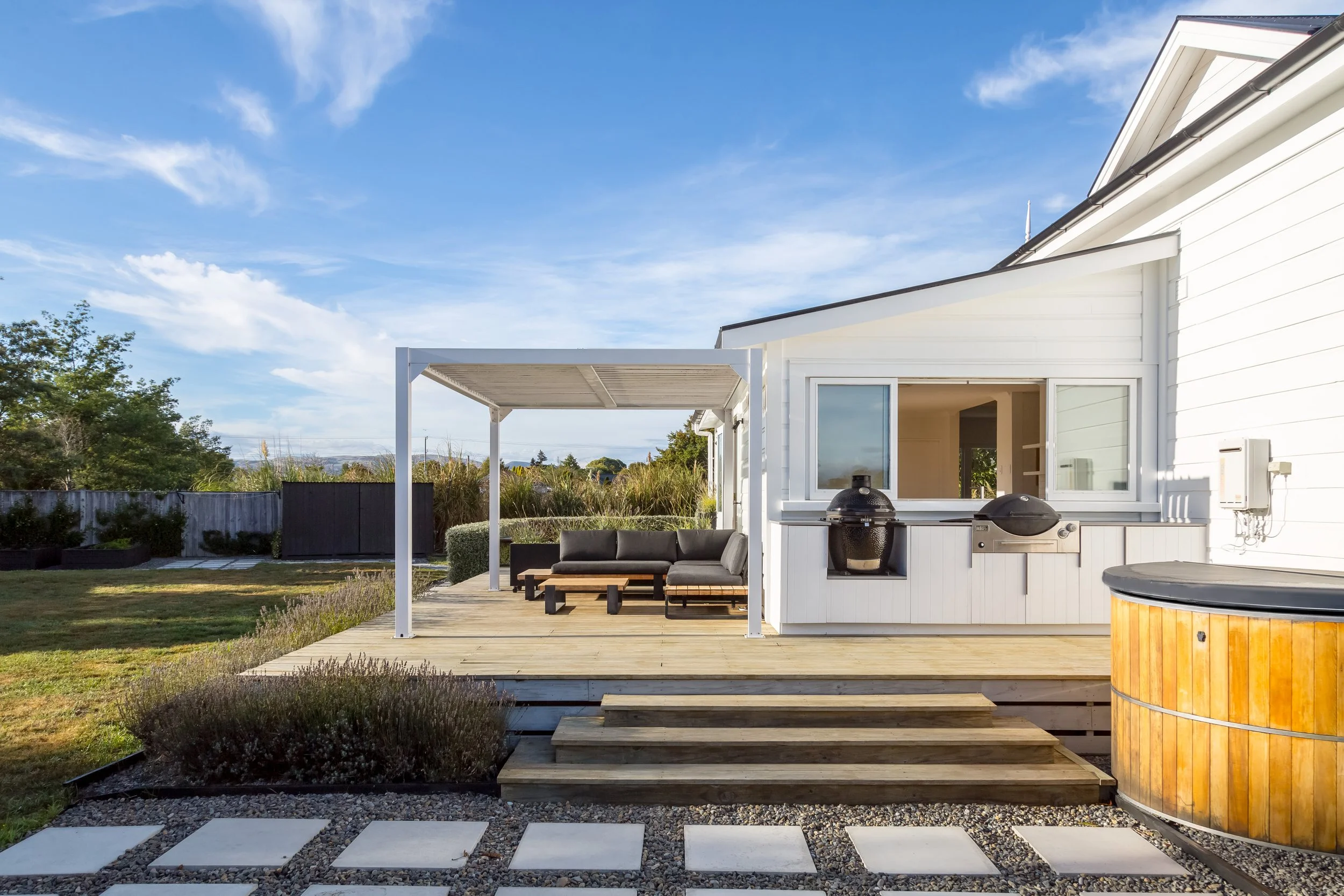 View of a large, charming white Victorian villa with a big sunny back deck area that has couches under a shaded pergola, along with an outdoor kitchen and spa, surrounded by well-kept lawns and trees under a blue sky.