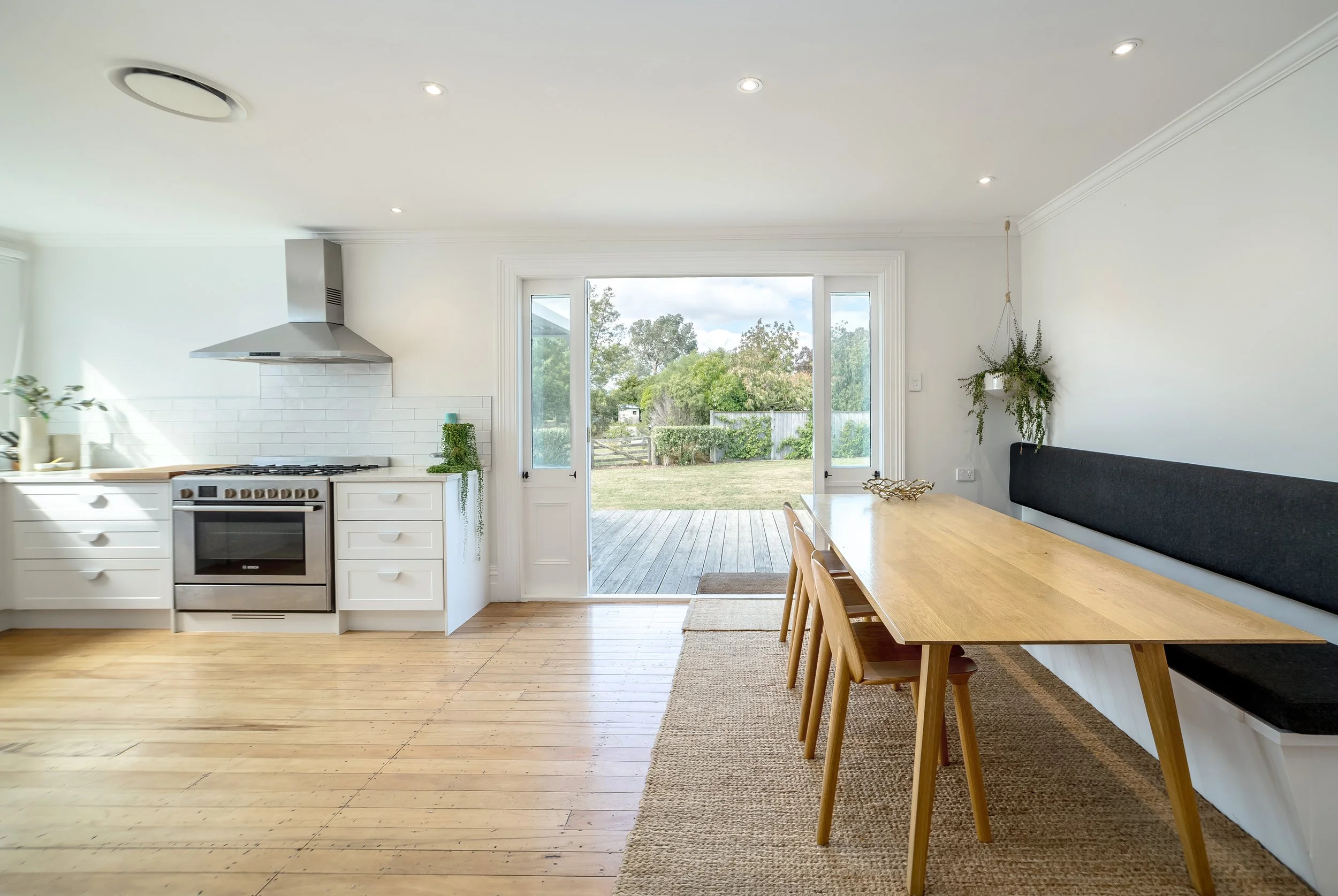 A bright white kitchen with sun streaming into it over light wood floors and wooden table, with chairs, a seat bench and french doors open, overlooking a wooden deck, green lawn and gardens. Great for weekend stays, group gatherings and holidays.