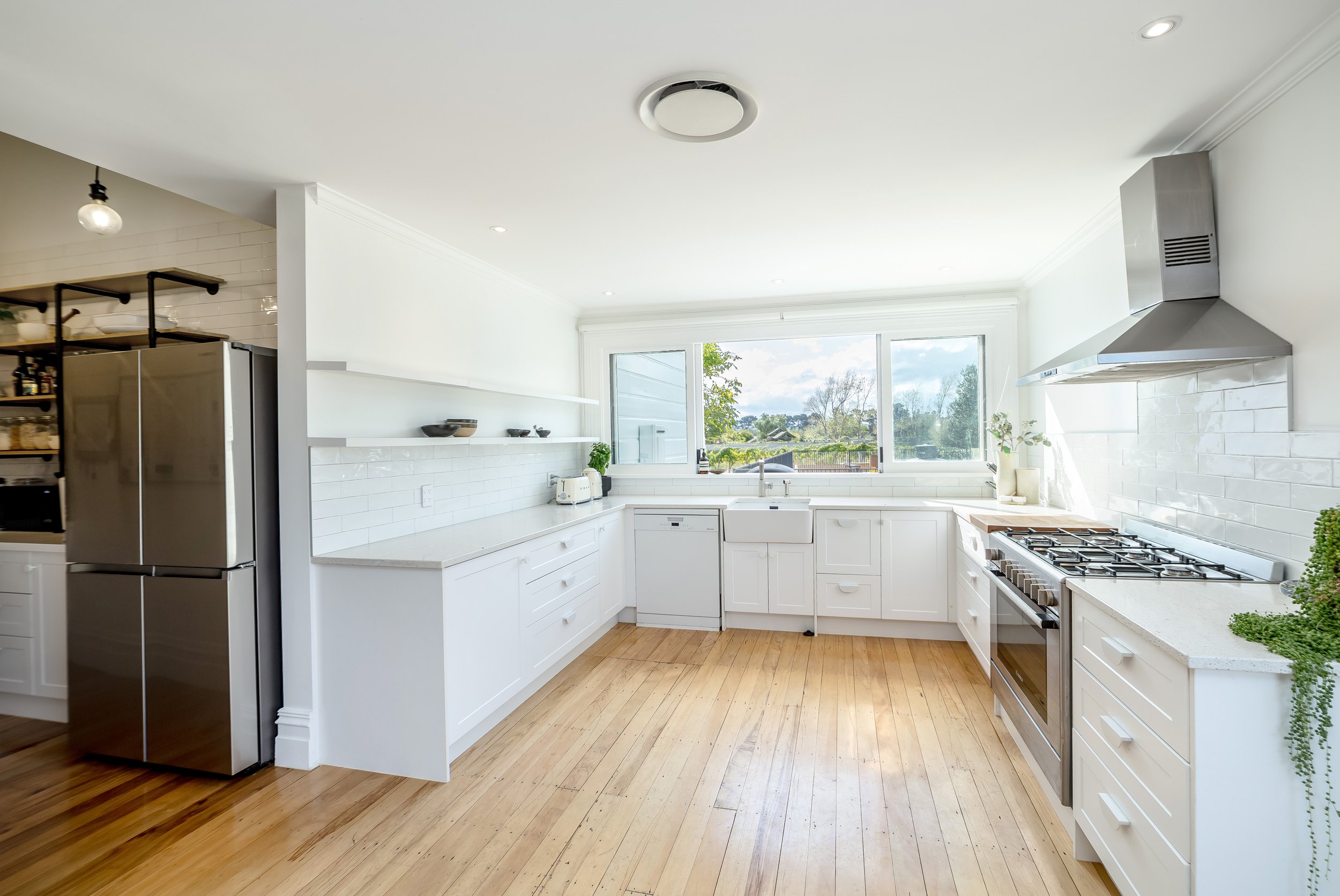 A contemporary bright white kitchen with white cabinets and bench, a light wood floor, large windows and a pantry off to one side with a large fridge. Great for weekend stays, group gatherings, celebrations and holidays.