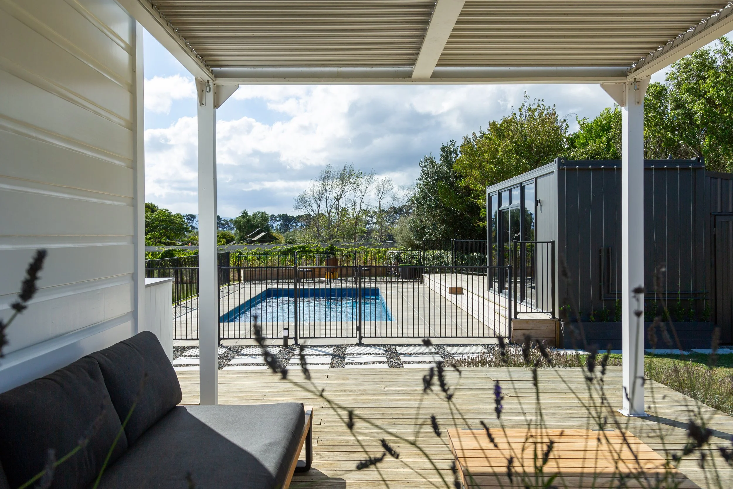 View from a covered pergola over a deck to a swimming pool, bordred by a pool house and trees under a blue sky dotted with clouds. Great for weekend stays, small weddings, post wedding events, group gatherings, celebrations and holidays.