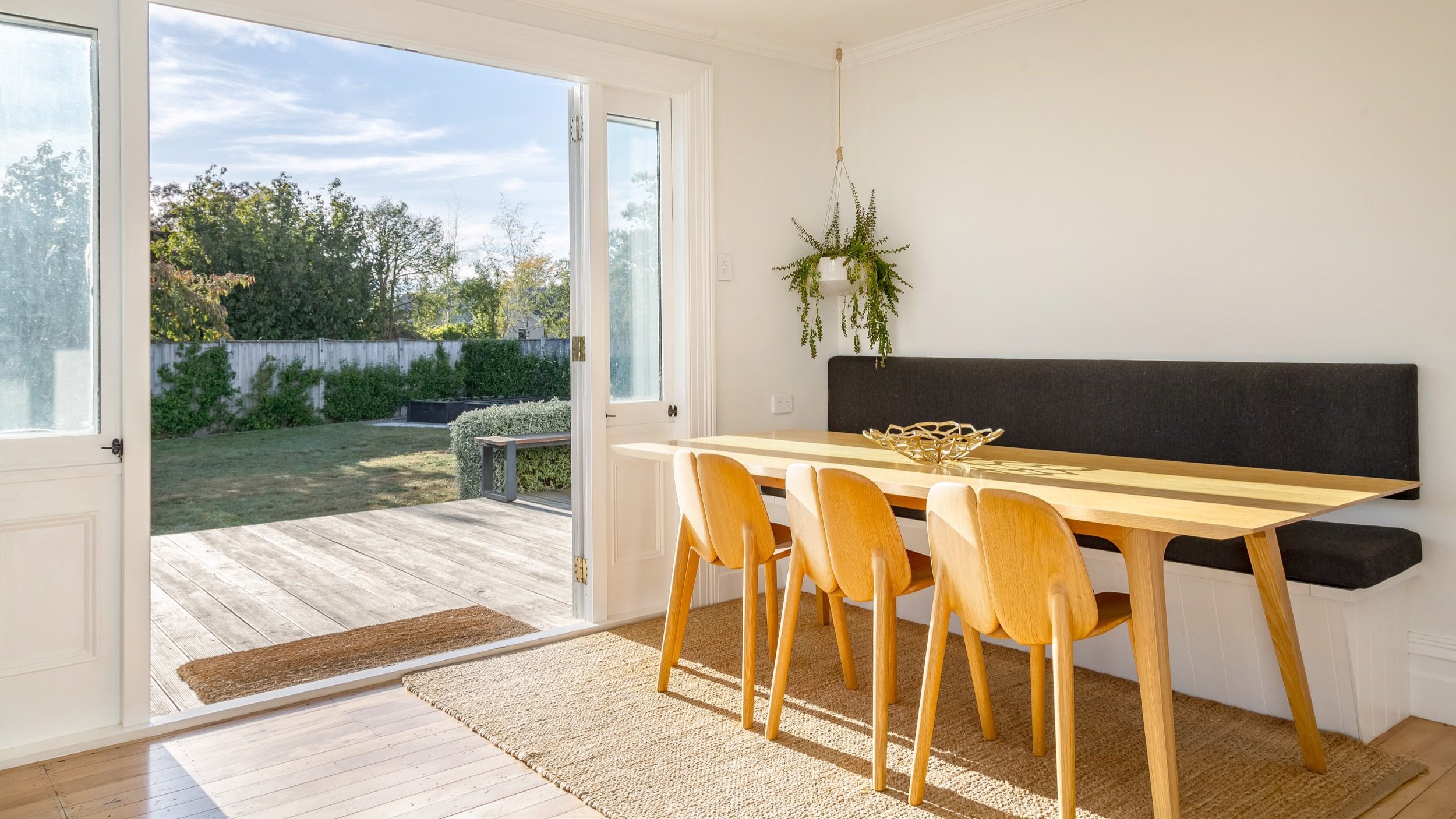 A dining area with warm sun streaming over a wooden table, overlooking a wooden deck, green lawn and gardens through an open door. Great for weekend stays, small weddings, post wedding events, group gatherings, celebrations and holidays.