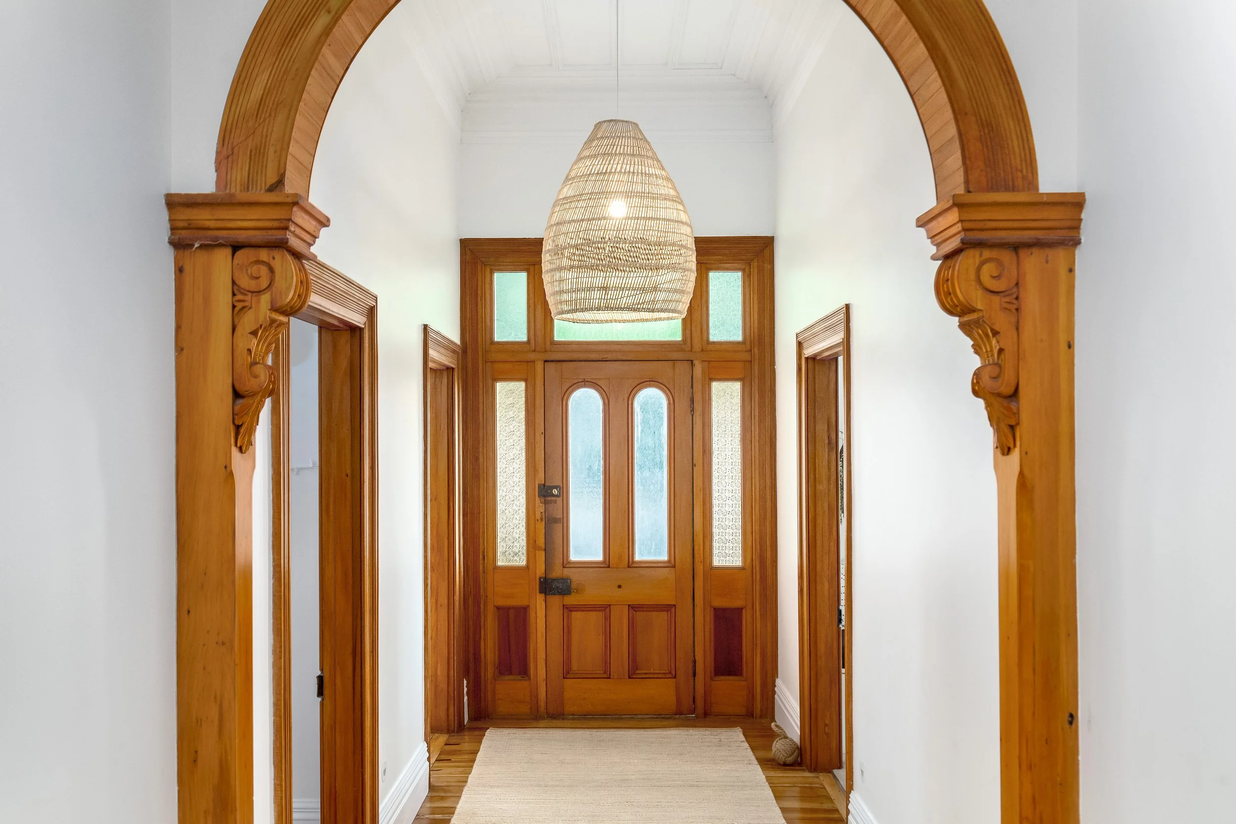 A large airy hall with a classic wooden front door, inset with frosted glass panels, and a modern woven pendant light, framed by an archway with ornate woodwork, and a warm, beach style rug on a light wooden floor.