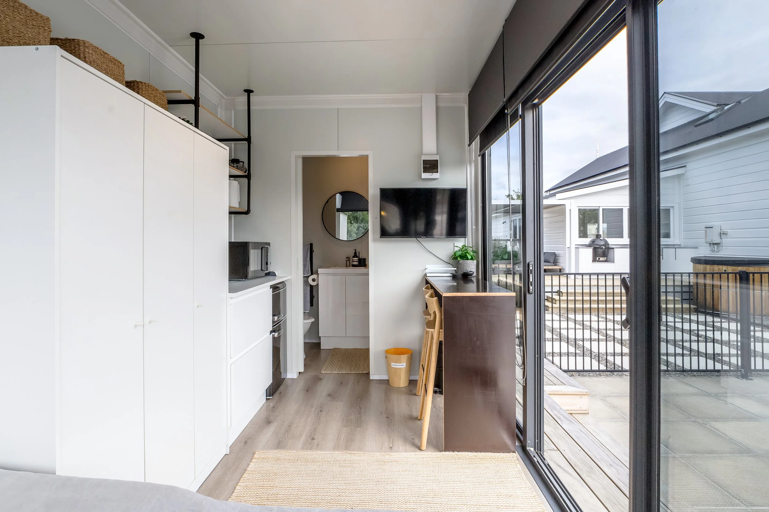 Interior view of the modern pool house living space with white cabinets, a microwave, TV, table with wooden stools, and a sliding glass door opening to a deck. Great for weekend stays, group gatherings, celebrations and holidays.