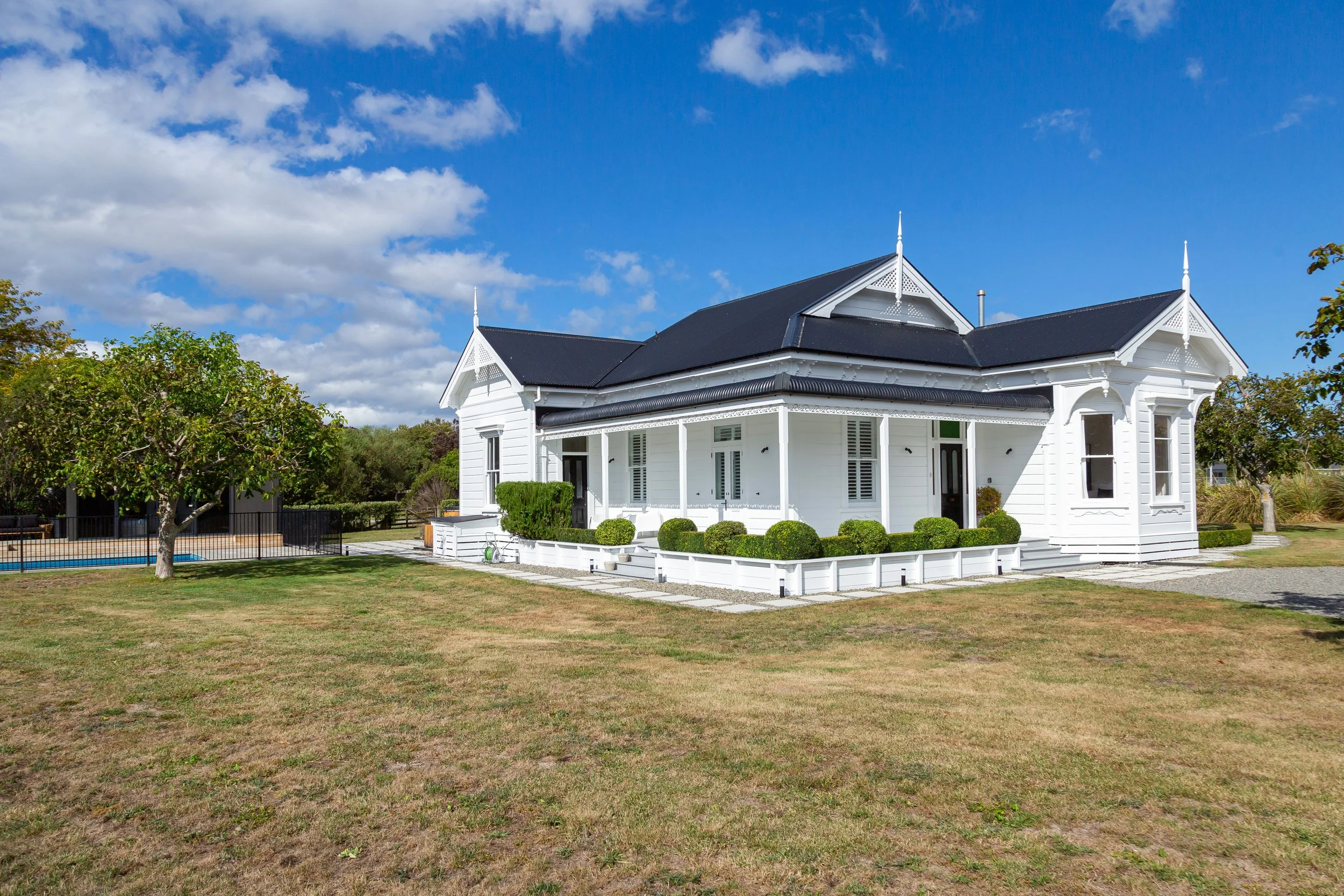 A large, charming white Victorian villa surrounded by well-kept lawns, topiary and trees under a blue sky dotted with a few white clouds. Great for weekend stays, small weddings, post wedding events, group gatherings, celebrations and holidays.