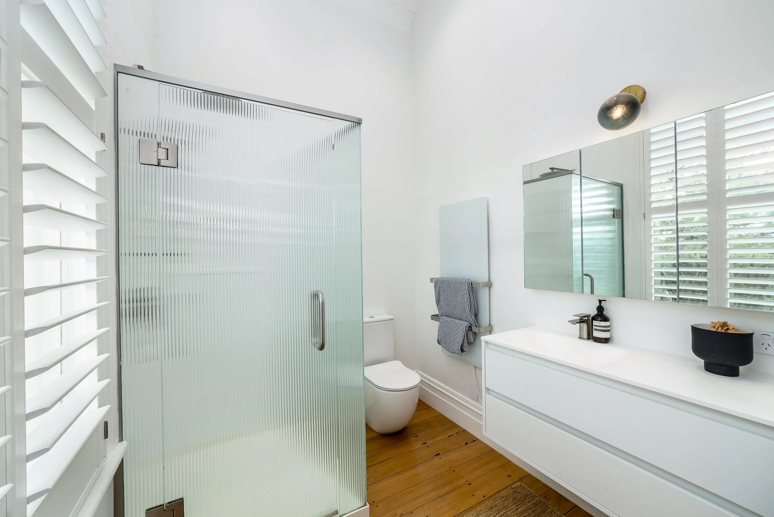 Light and airy modern bathroom with white walls, wooden floor, fluted glass shower, large mirror cabinet above a white vanity and white wooden blinds. Great for weekend stays, group gatherings, celebrations and holidays.