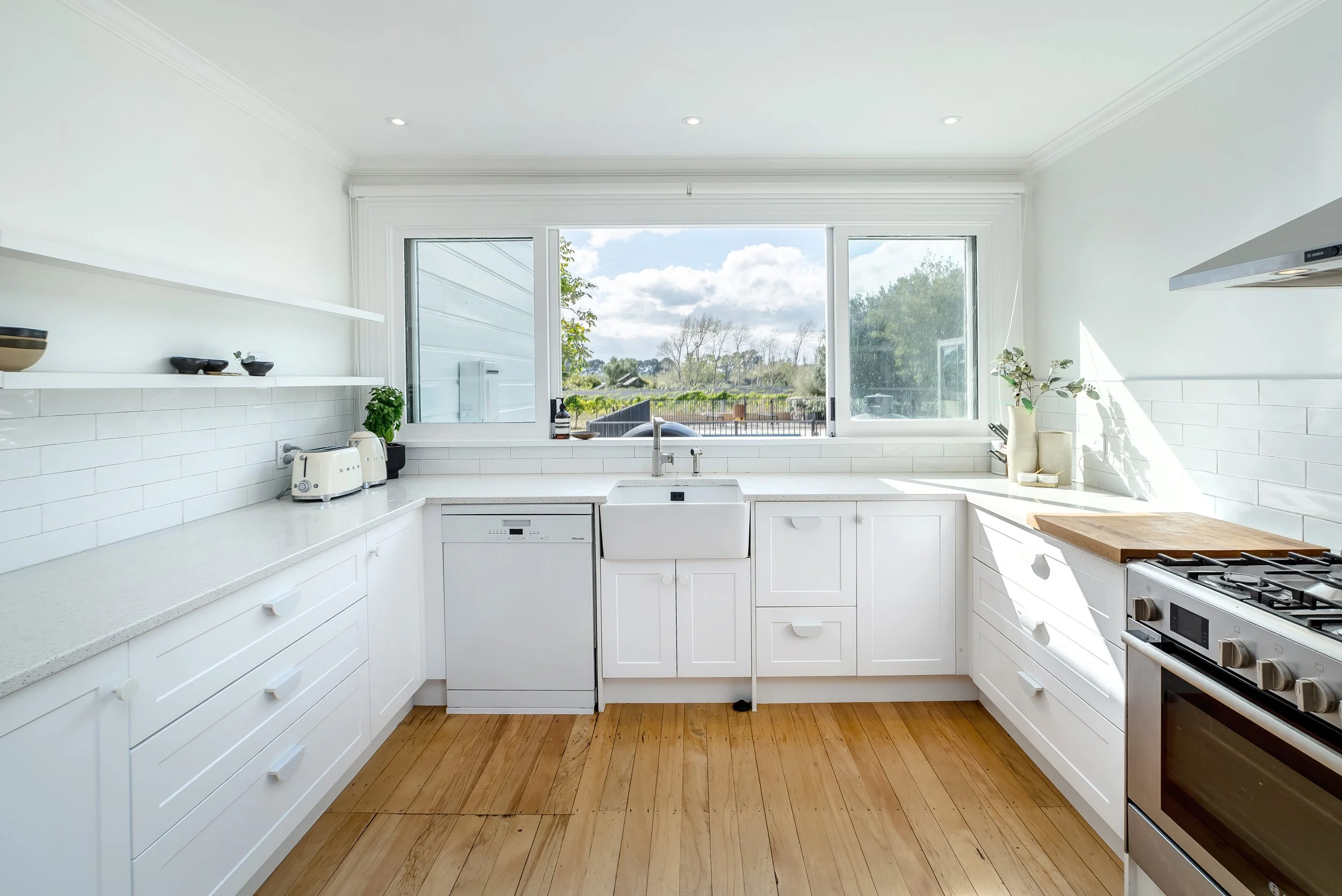 A contemporary bright white kitchen with white cabinets and bench, white subway tiles, a six burner oven, dishwasher and a light wood floor and large windows that look out to a vineyard scene.