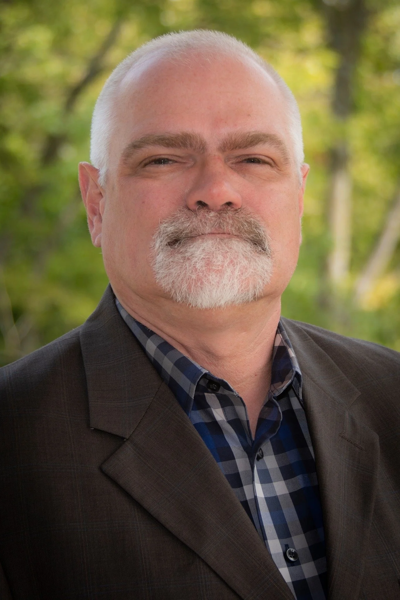 Close-up of a middle-aged man with a white beard and mustache, dressed in a plaid shirt and dark blazer, standing outdoors with blurred green trees in the background. Dan Hostert.