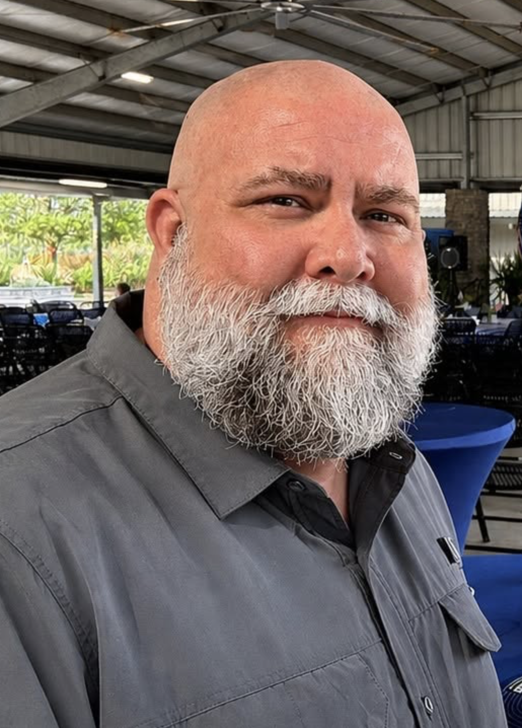 A middle-aged man with a bald head and a long white beard, wearing a gray shirt, smiling at the camera inside a covered outdoor space with tables and chairs.