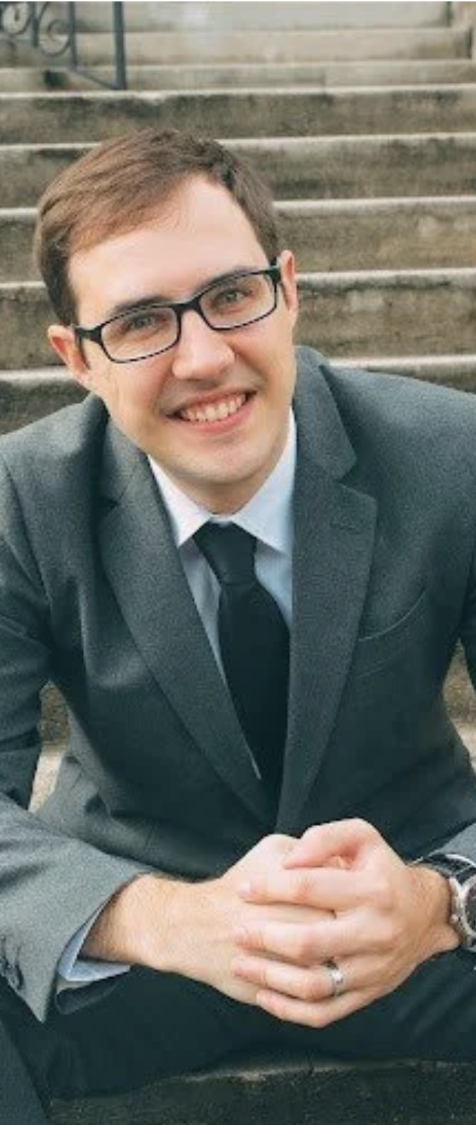 A young man with short brown hair, glasses, and light skin, smiling and wearing a gray suit with a white shirt and black tie, sitting on outdoor stone stairs.
