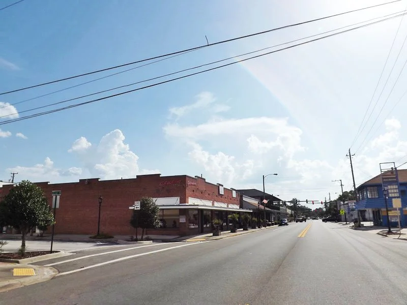 A street scene in a small town with brick buildings, a blue building on the right, trees, utility poles, and a partly cloudy sky with the sun shining.