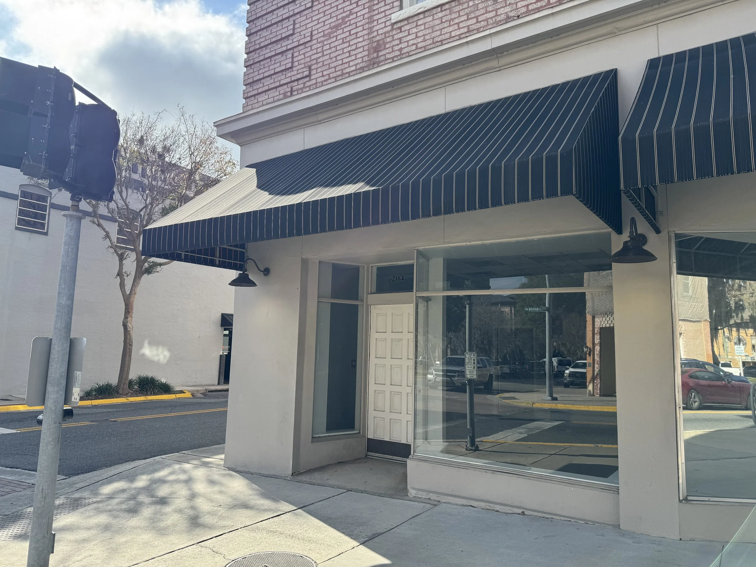 Empty storefront with large glass windows and a white door, blue striped awning, sidewalk, and street with parked cars in the background.