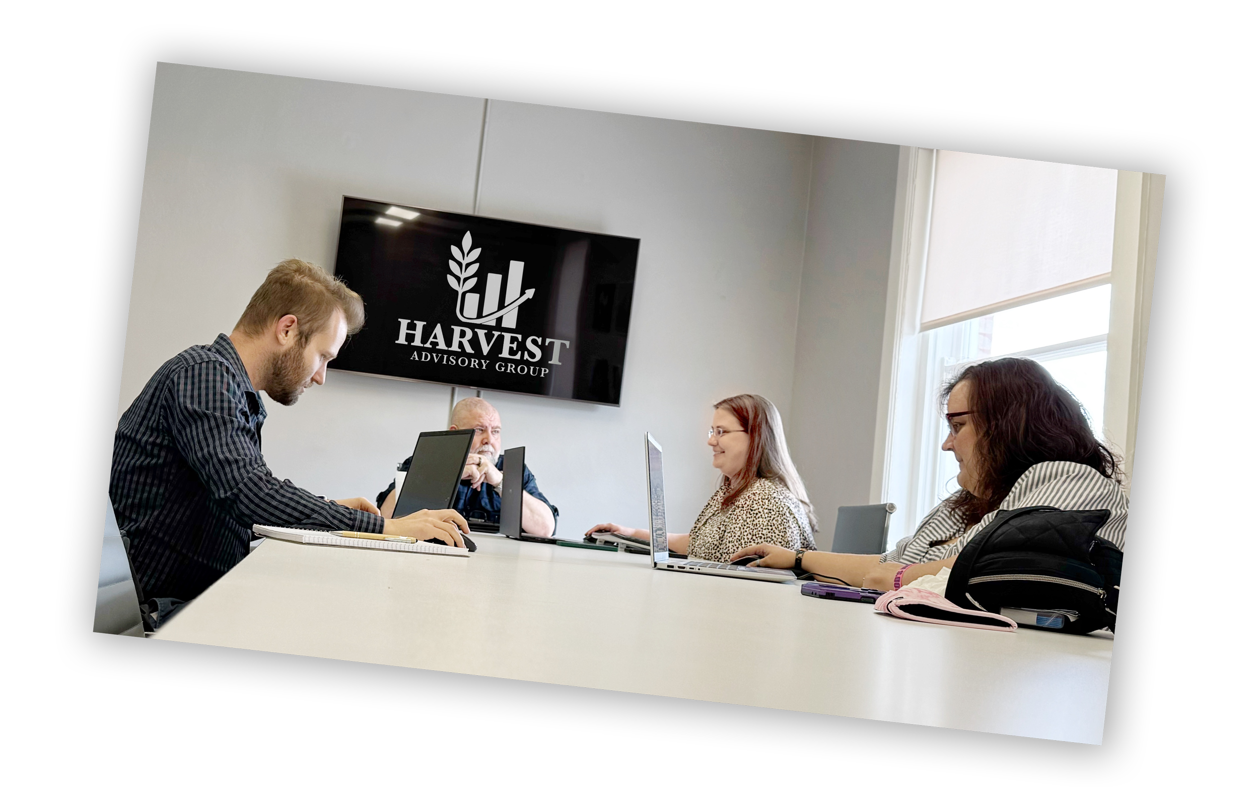 People sitting at a conference table with laptops and notebooks, in a meeting room with a large window and a screen displaying the Harvest Advisory Group logo.