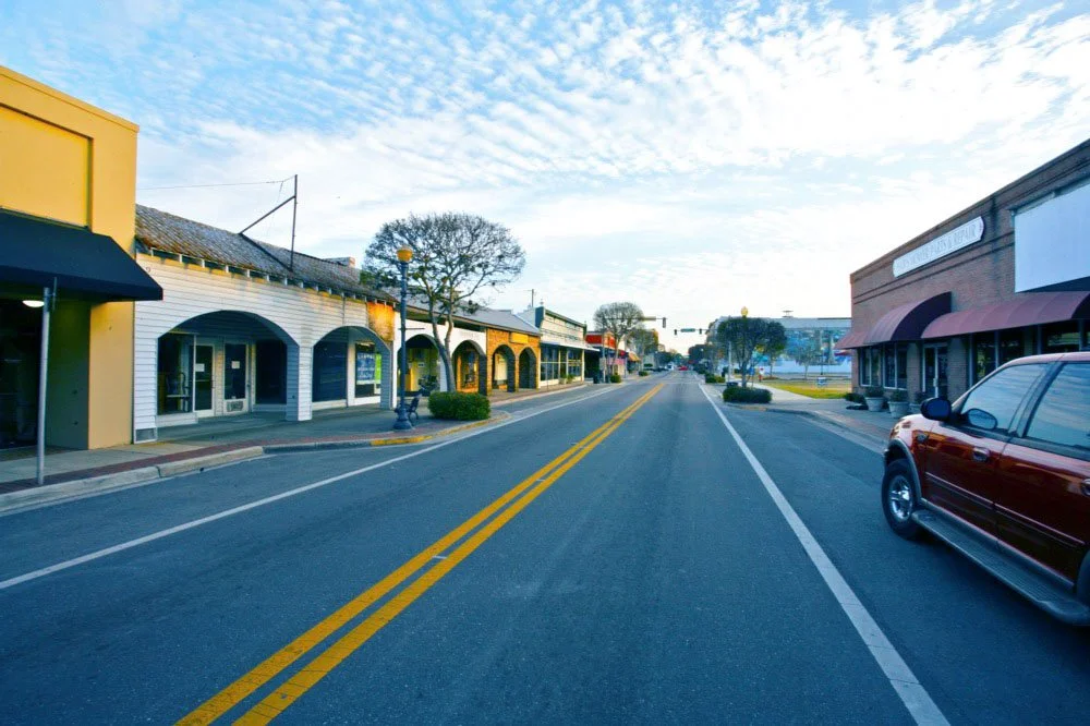 Empty downtown street with storefronts on both sides, a parked red SUV on the right, a tree, and clear blue sky with some clouds.