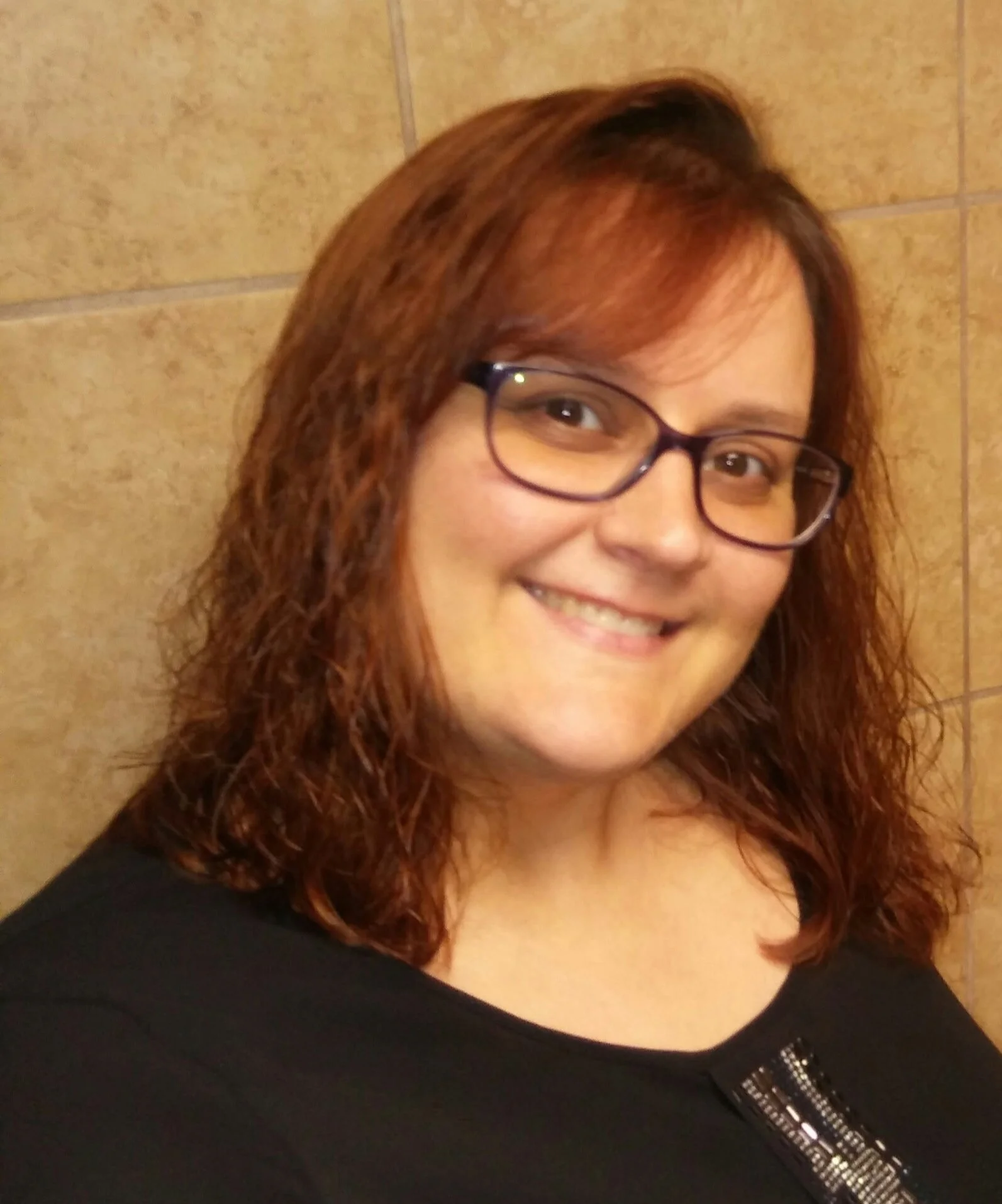 Close-up of a woman with reddish-brown hair, wearing glasses, smiling, dressed in black, against a beige tiled background.