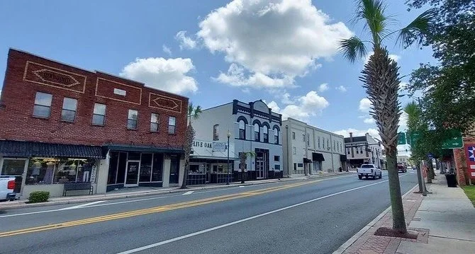 Street view of small town with brick and painted storefronts, palm trees, parked cars, and blue sky with clouds.