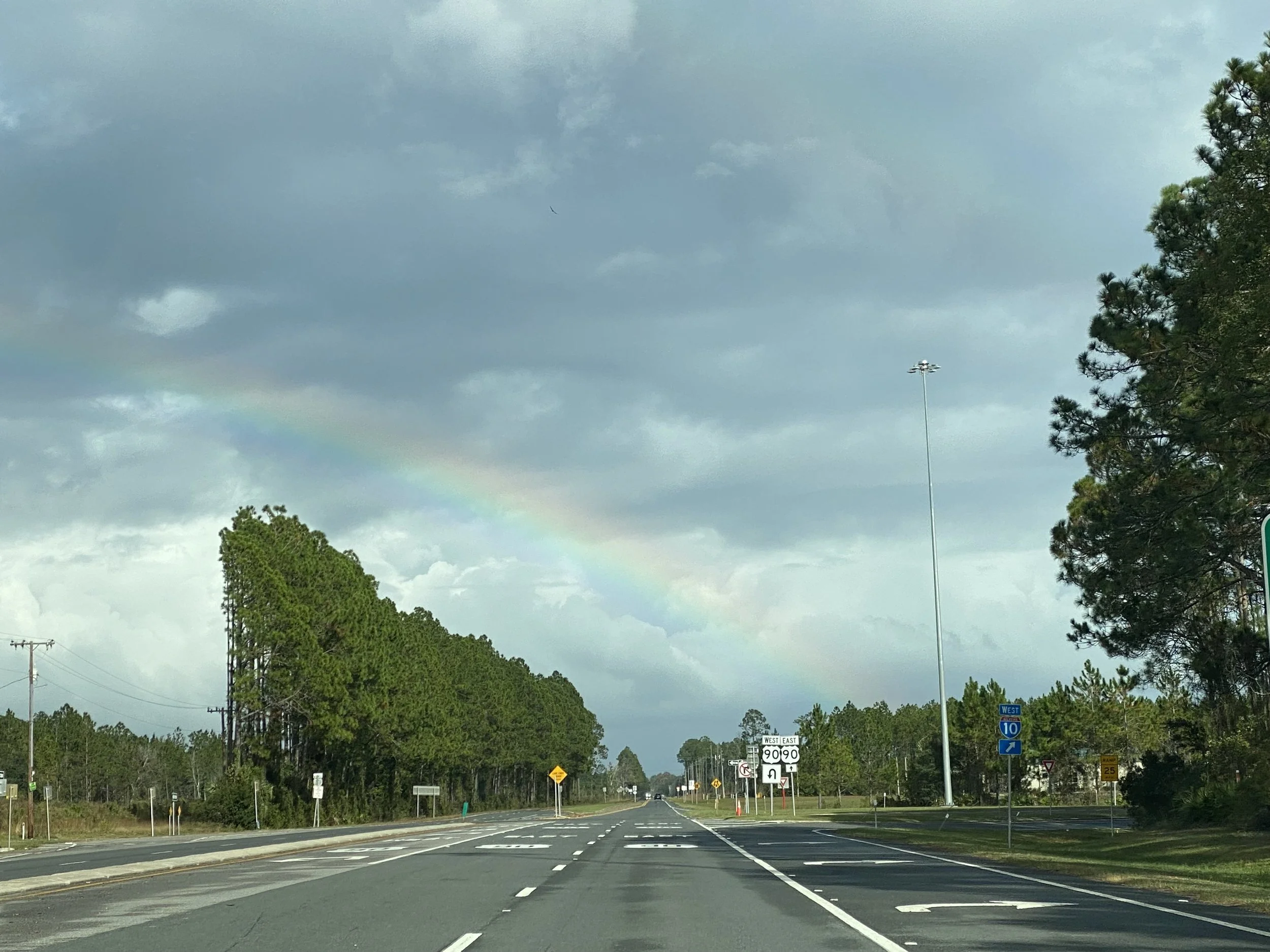 A rural highway scene with a rainbow in a cloudy sky, green trees on both sides, and road signs including directions for Interstate 10 and U.S. Route 90.