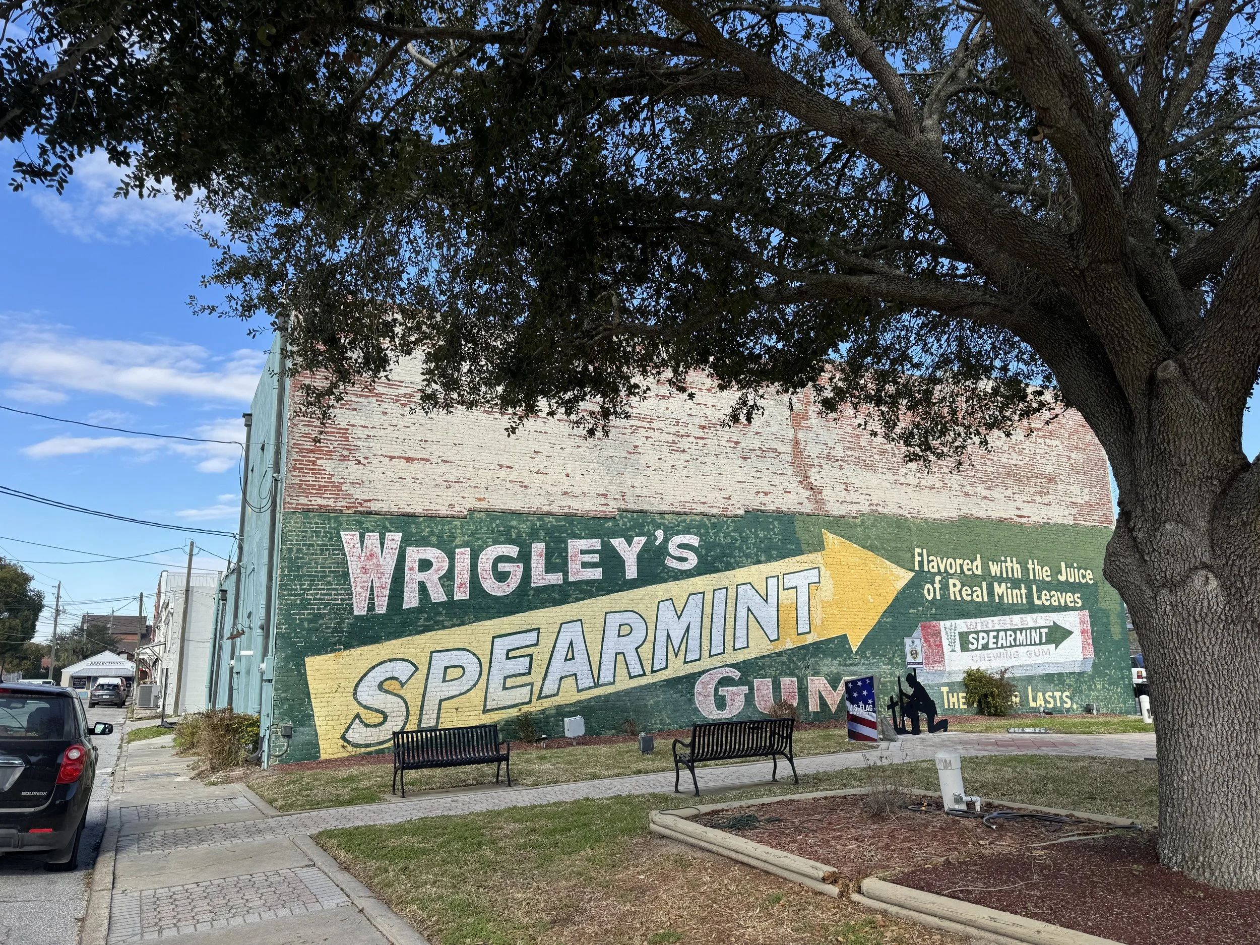 A colorful vintage advertisement painted on a brick wall for Wrigley's Spearmint Gum, with a large arrow and the text "Flavored with the Juice of Real Mint Leaves". There are benches, trees, and parked cars nearby. Located in downtown Live Oak, FL.