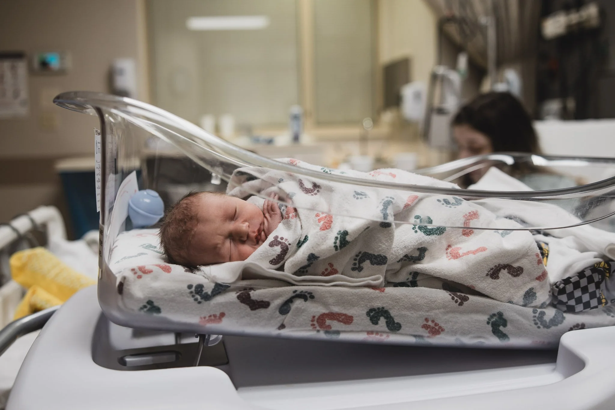 A newborn baby sleeps in his hospital bed as his mom rests in the background.