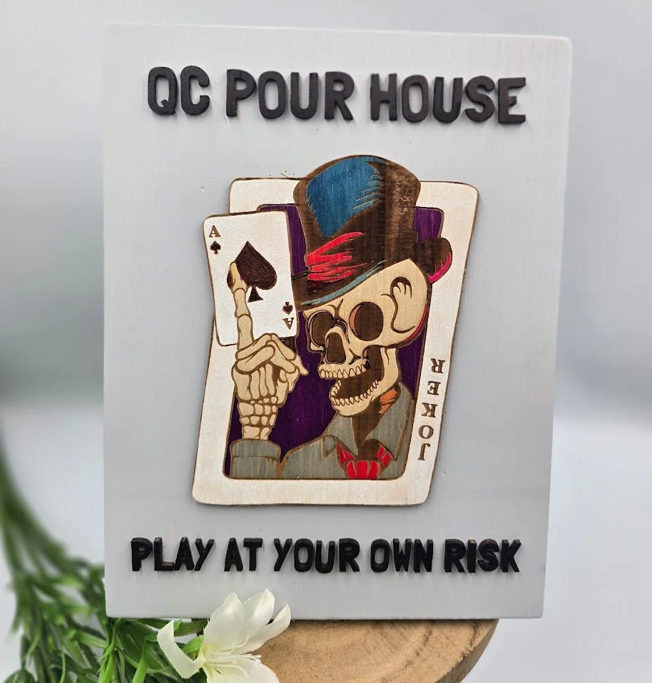 Sign with a cartoon skeleton wearing a hat and holding a playing card, with text stating "QC POUR HOUSE" at the top and "PLAY AT YOUR OWN RISK" at the bottom.