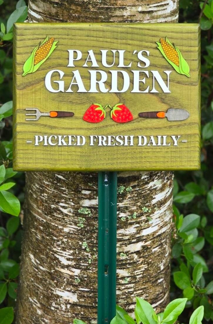 A wooden sign reading 'Paul's Garden, Picked Fresh Daily,' with illustrations of strawberries and gardening tools, leaning on a tree trunk surrounded by green leaves.