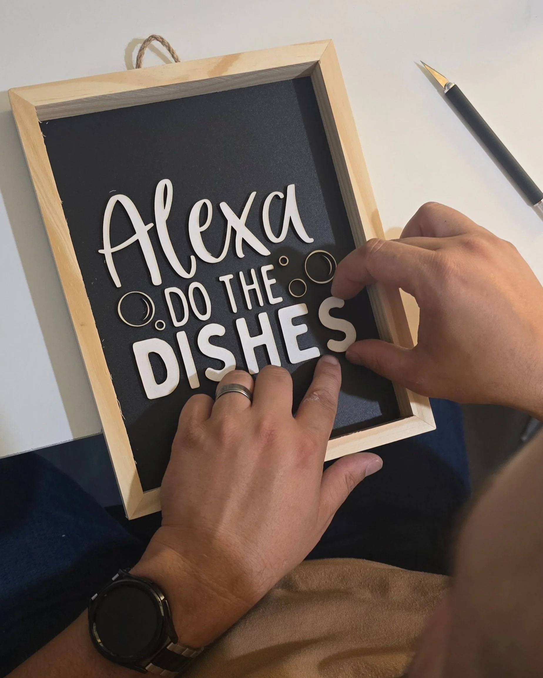 Person arranging white letters on a blackboard with a wooden frame, spelling out 'Alexa, DO THE DISHES,' with a black marker pen placed on the table nearby.