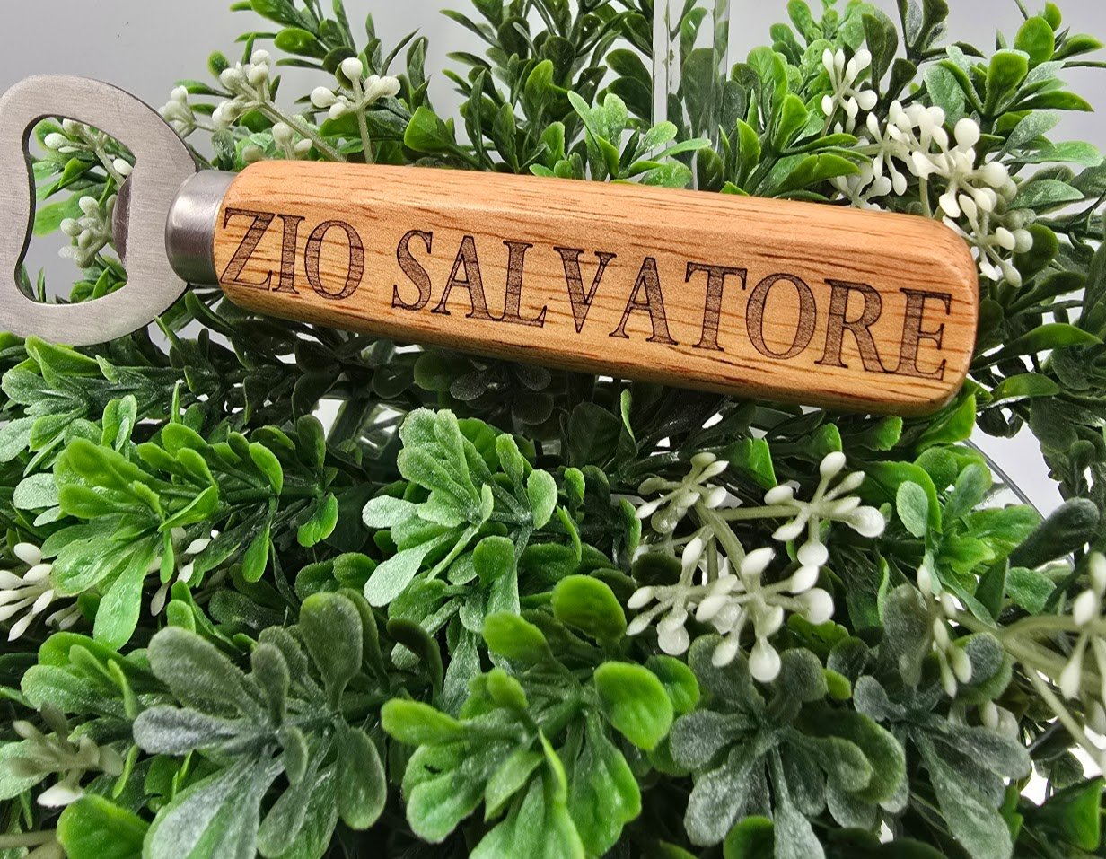 A wine bottle opener with a wooden handle engraved with 'ZIO SALVATORE' resting on green foliage with white small flowers.